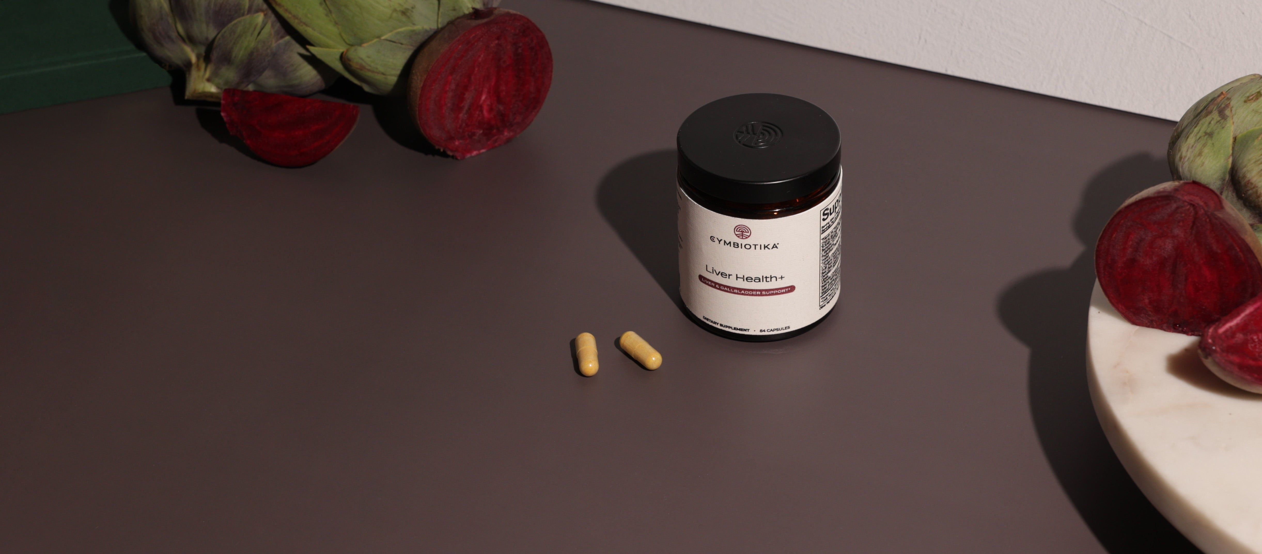 Cymbiotika Liver Health+ jar with two capsules on a dark tabletop beside beets