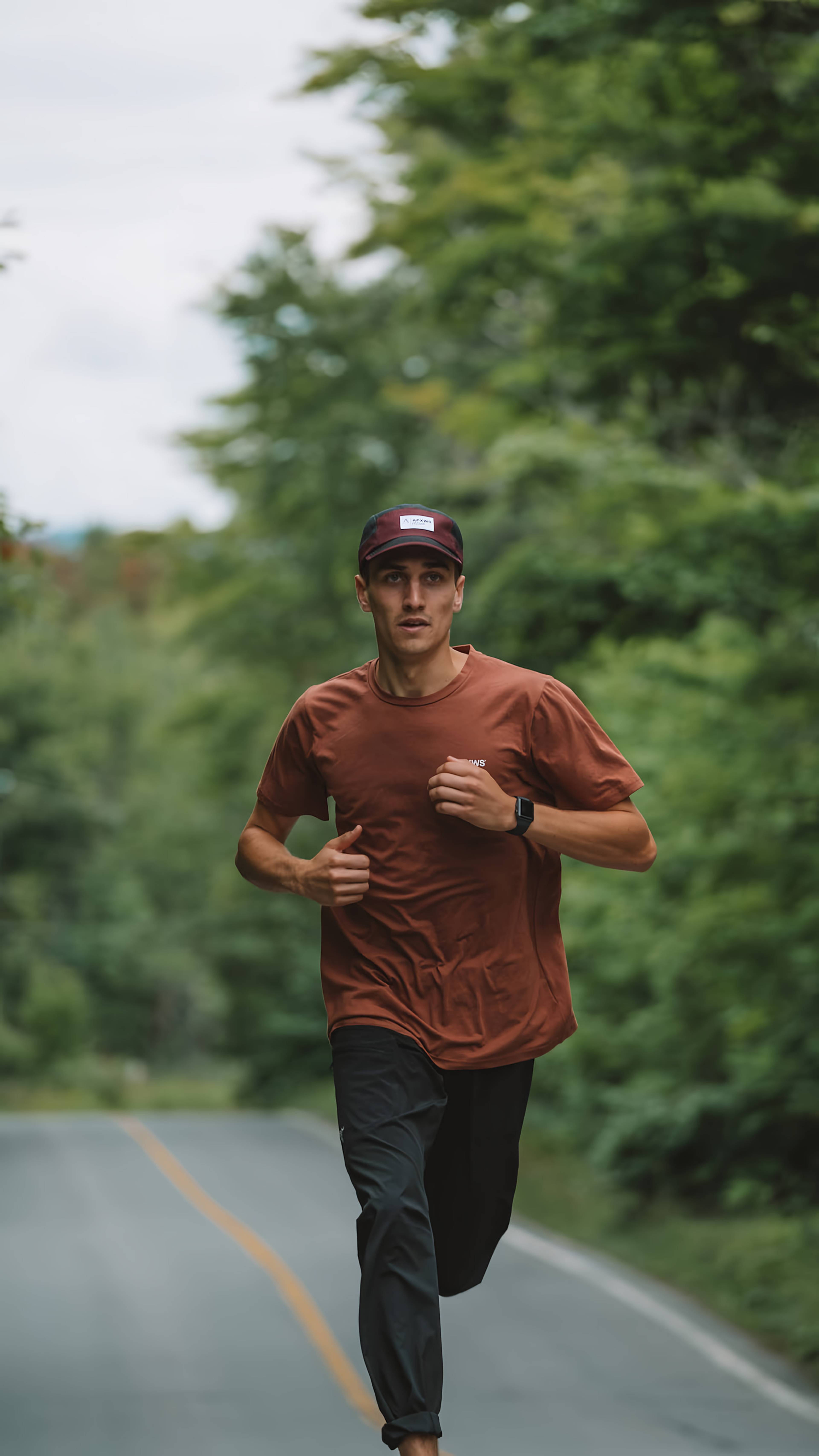 Man in a rust-colored shirt and cap running on a tree-lined road.