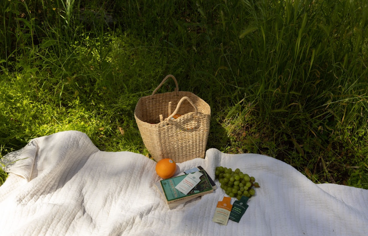 Woven basket, orange, green grapes, books, and small supplement packets on a white blanket in grass.