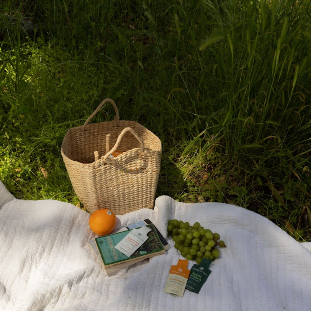 Woven basket, orange, green grapes, books, and small supplement packets on a white blanket in grass.