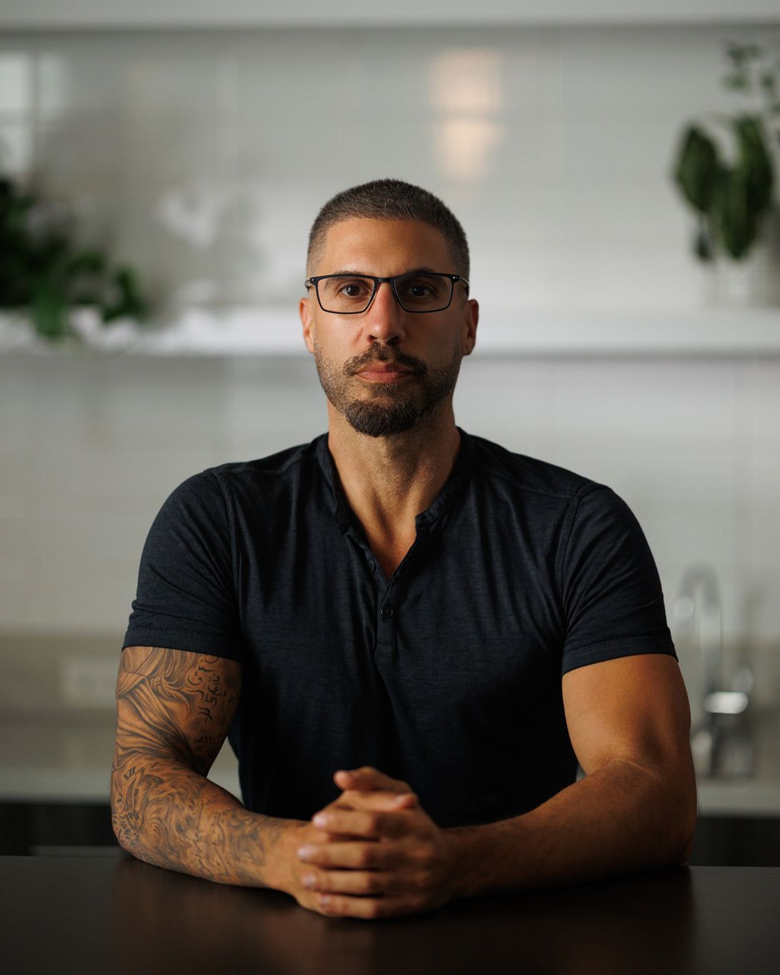 A bespectacled man with a beard and tattooed arm sits at a table with his hands clasped.