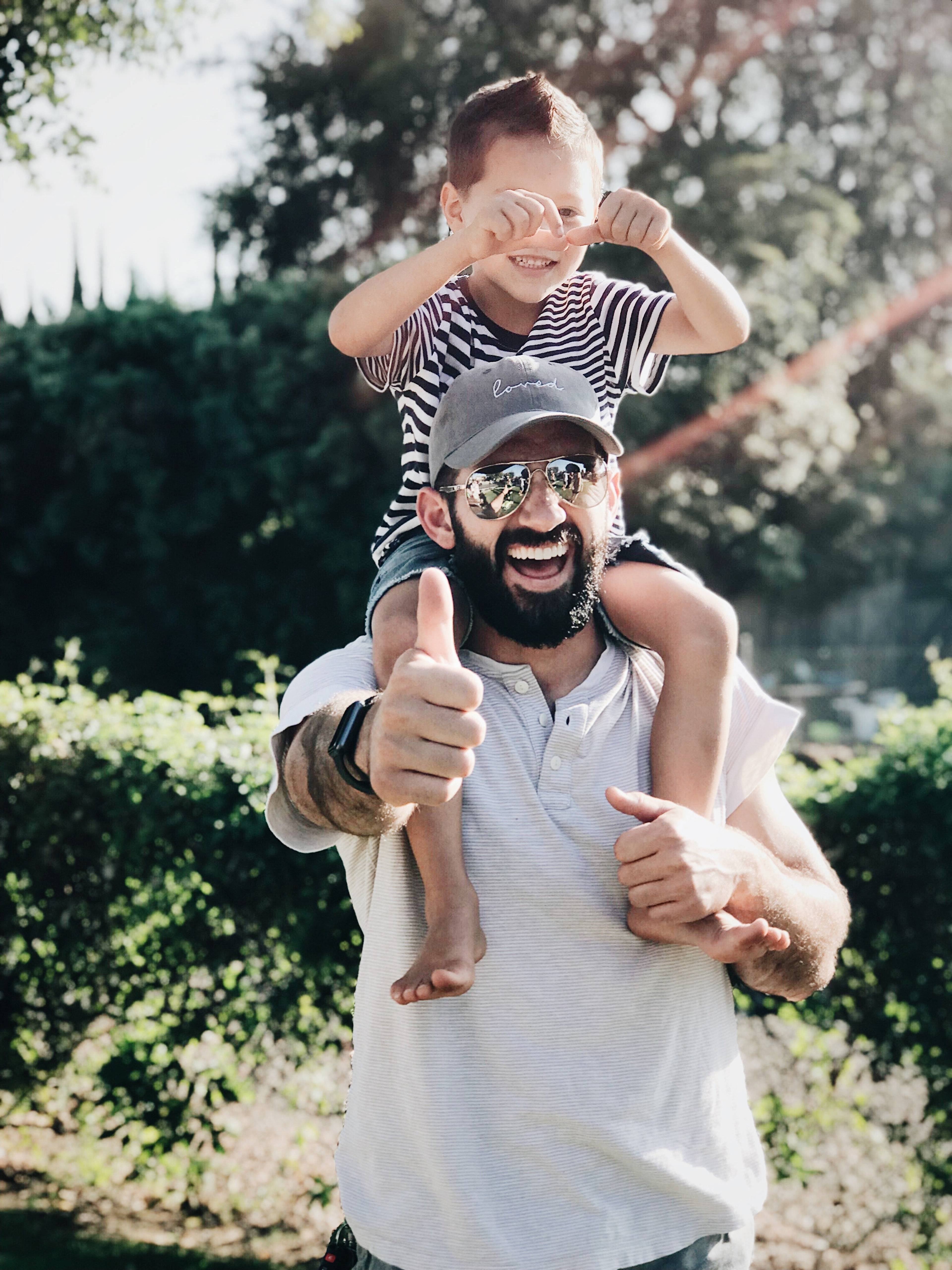 Smiling man gives a thumbs-up while carrying a child on his shoulders outdoors.
