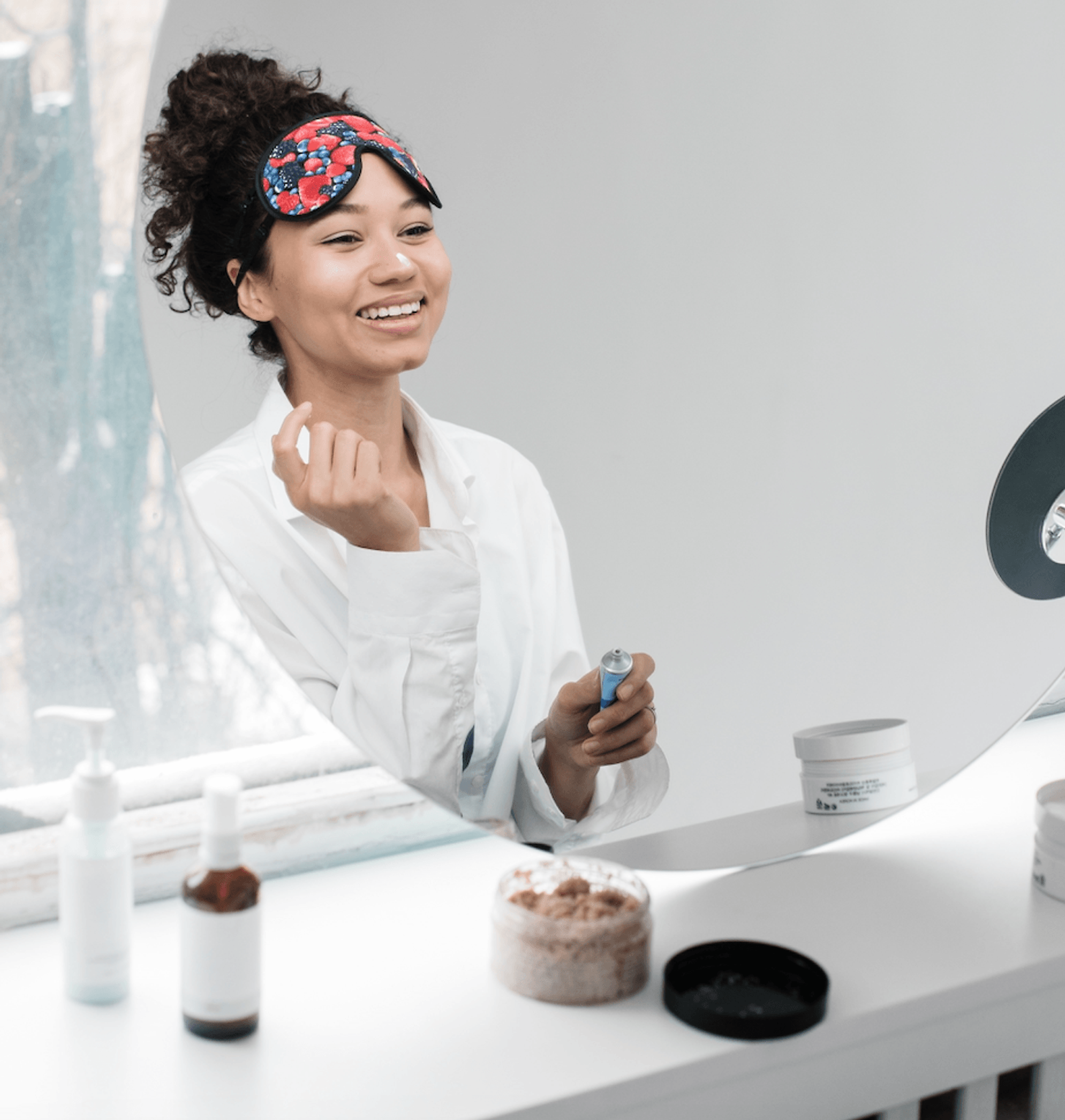 Smiling woman in a white shirt holds a skincare bottle at a vanity, wearing a colorful sleep mask on her forehead.
