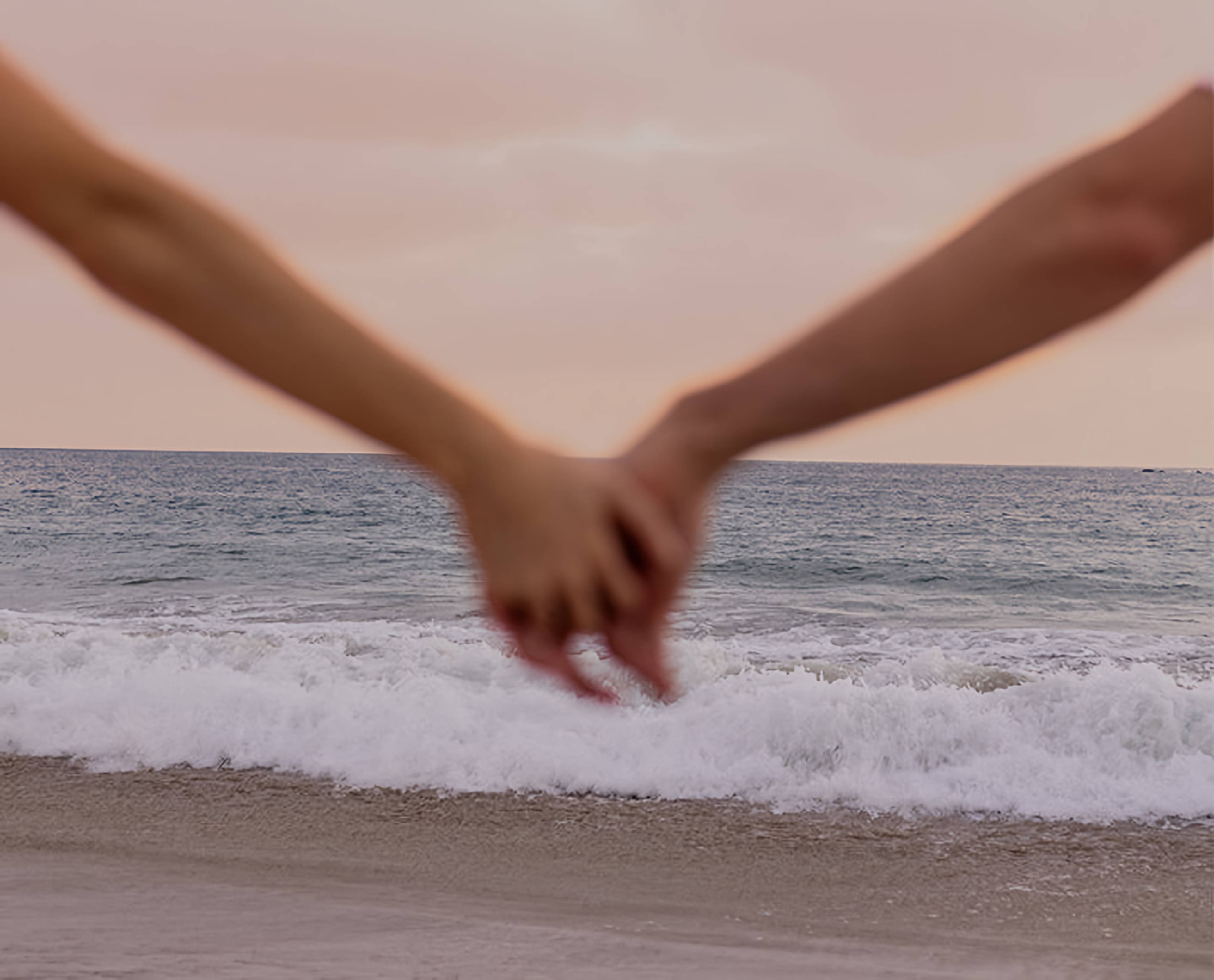 Two hands clasped above breaking waves on a beach.