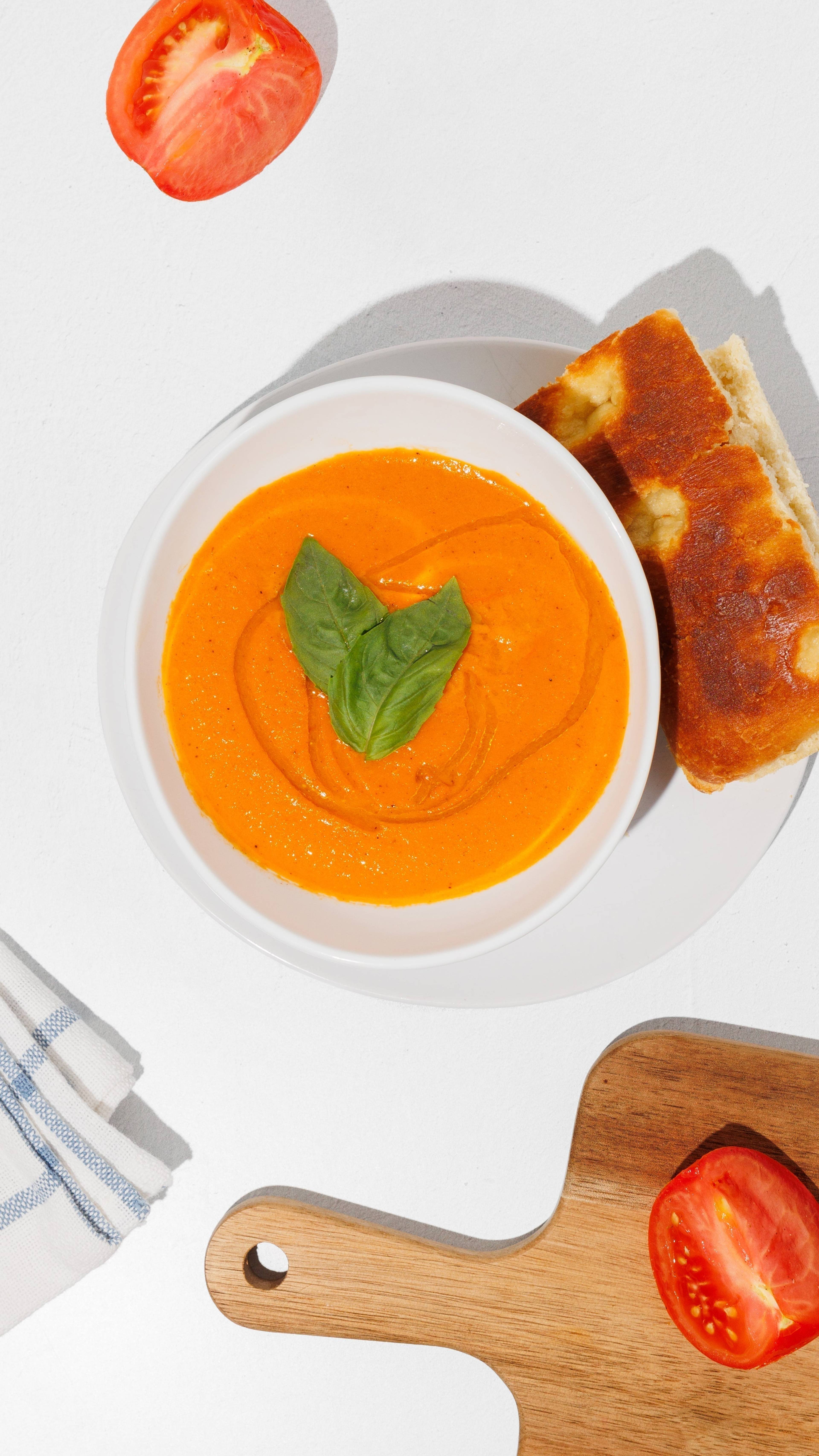 Bowl of tomato basil soup with a basil garnish, bread, tomato halves, a striped towel, and a wooden cutting board.