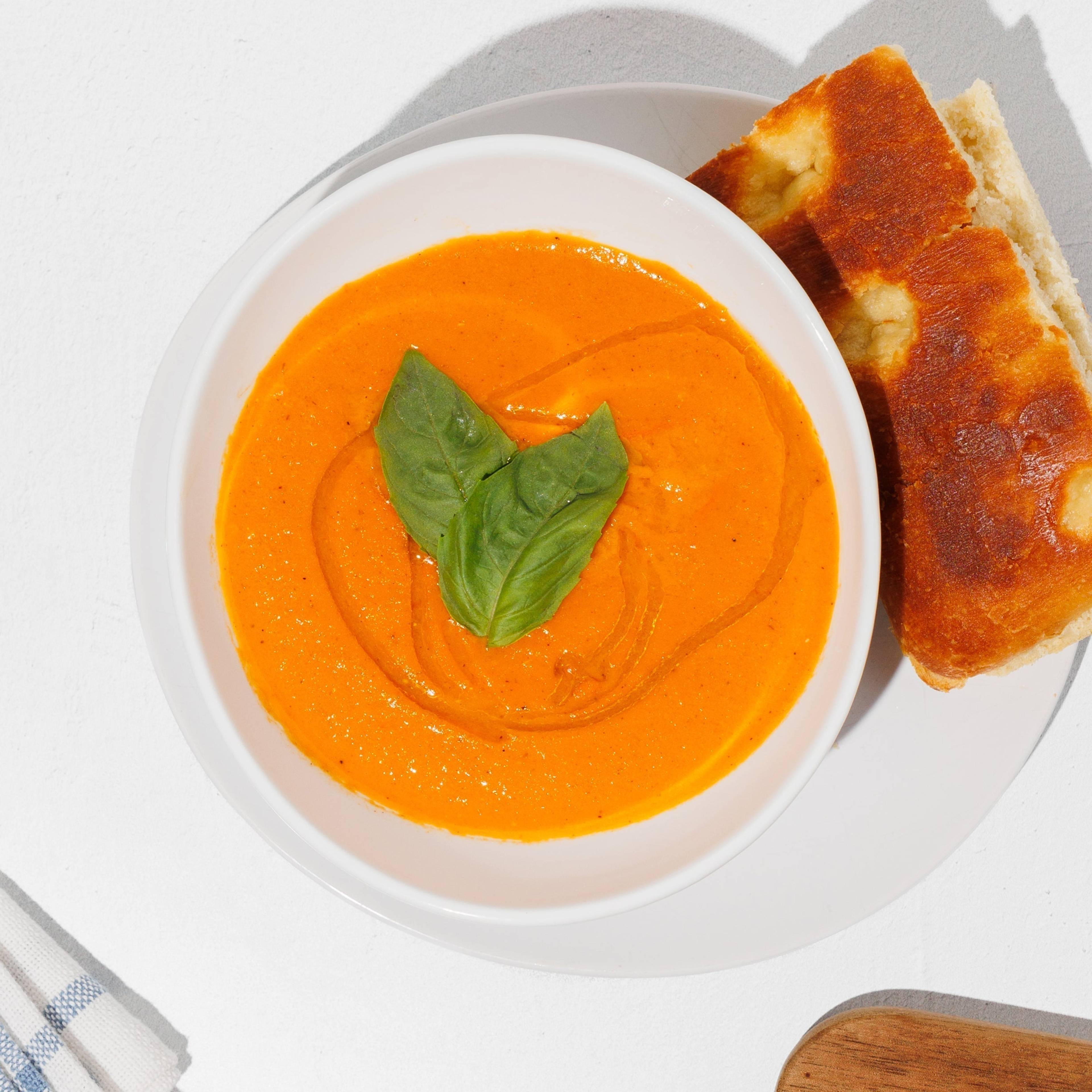 Bowl of tomato basil soup with a basil garnish, bread, tomato halves, a striped towel, and a wooden cutting board.