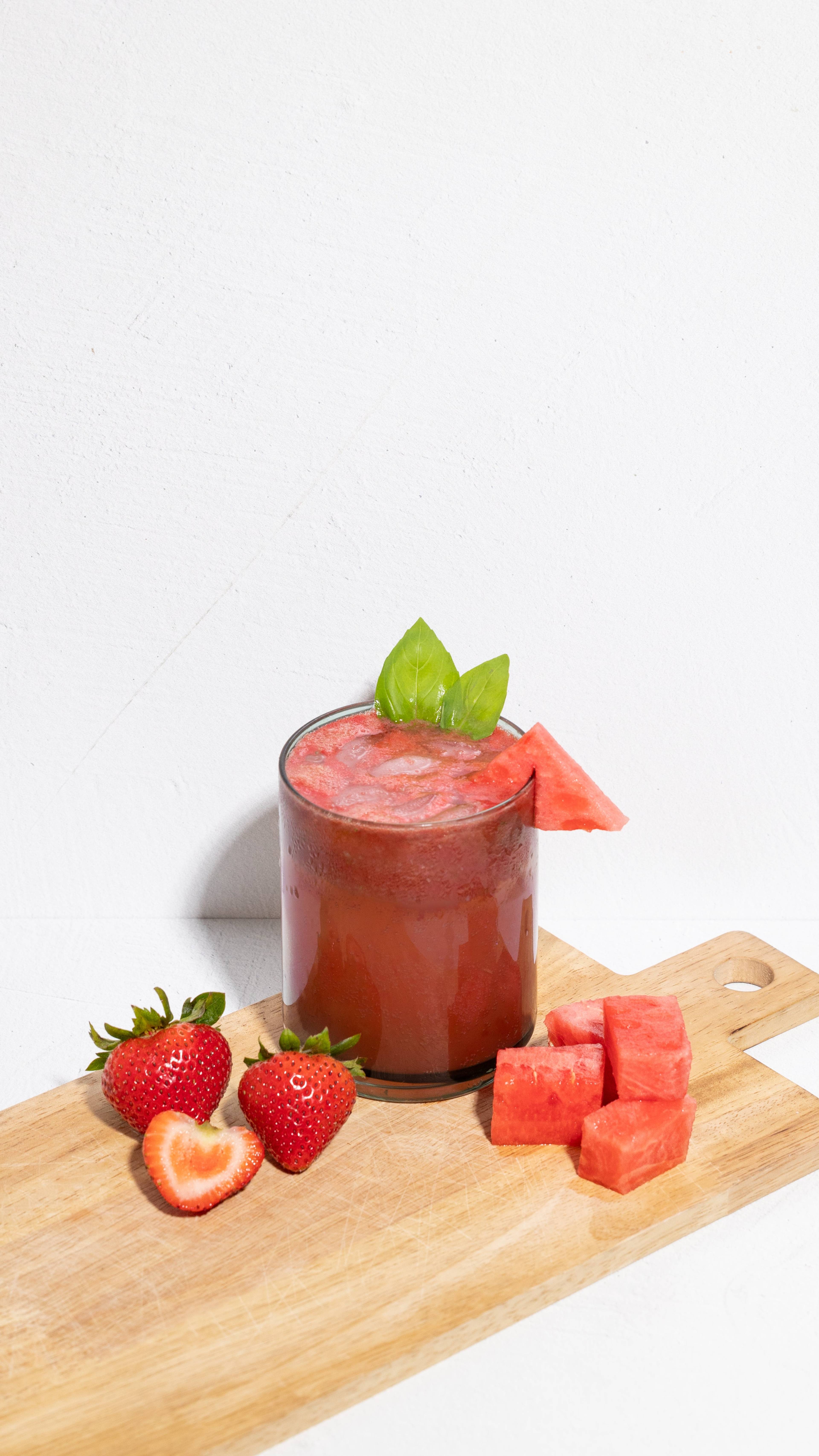 Glass of red iced juice topped with green leaves and a watermelon wedge, with strawberries and watermelon cubes on a board.