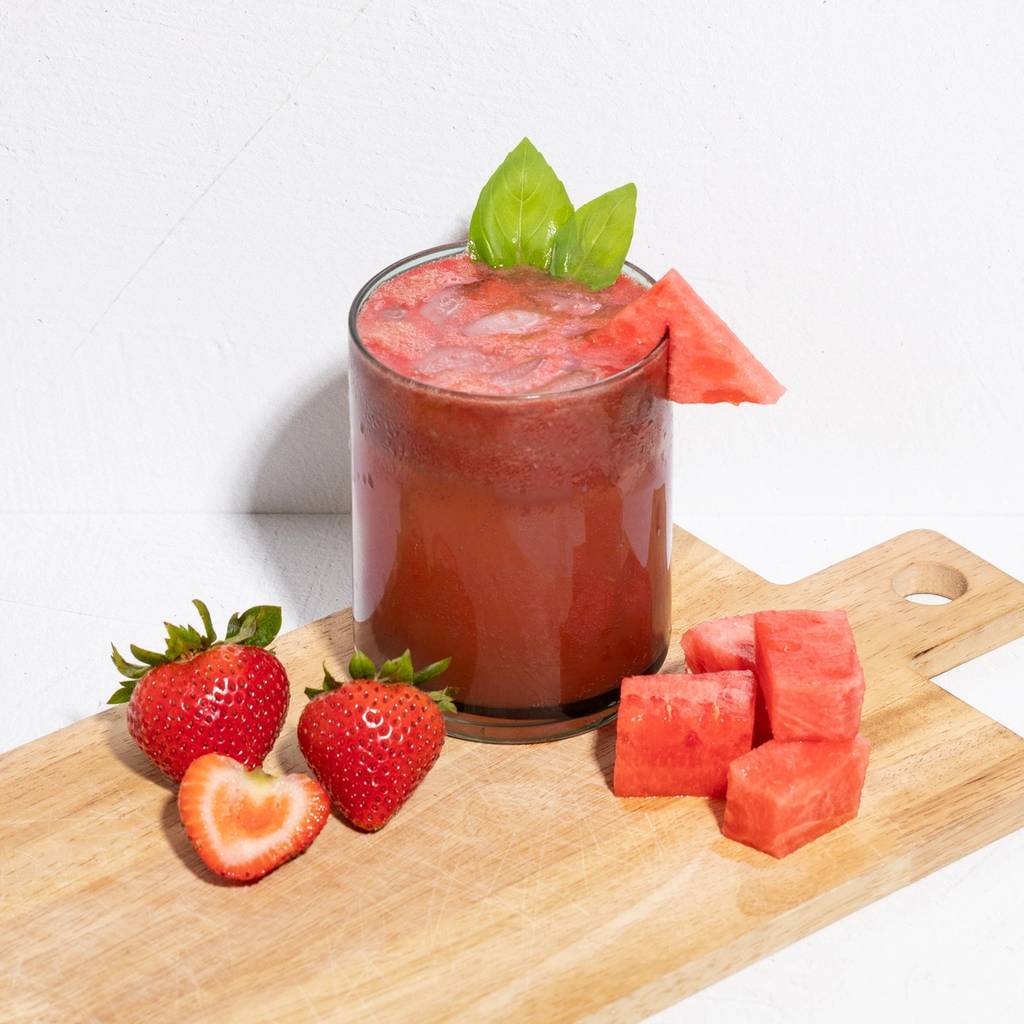 Glass of red iced juice topped with green leaves and a watermelon wedge, with strawberries and watermelon cubes on a board.