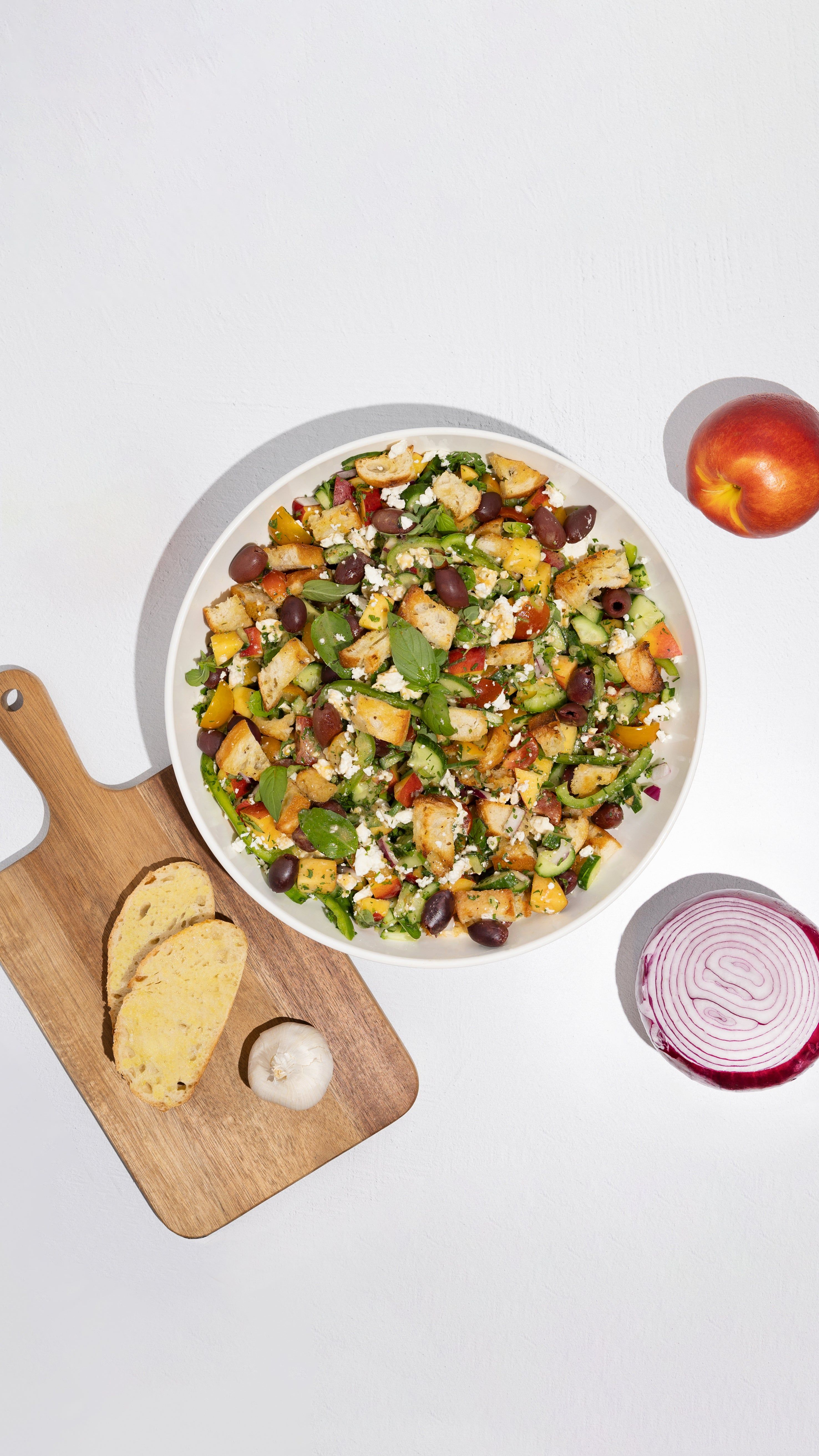 Overhead view of a bowl of Greek-style salad with bread, garlic, red onion, and a peach.