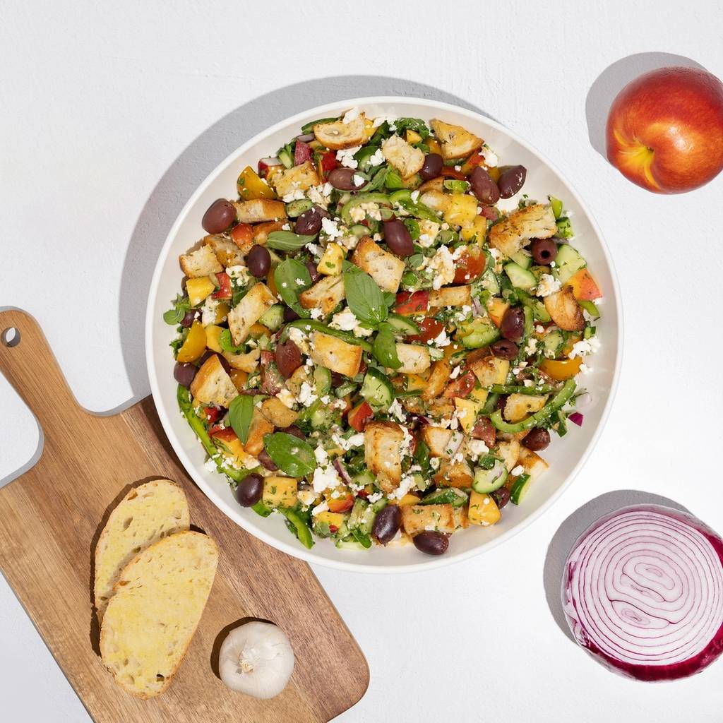 Overhead view of a bowl of Greek-style salad with bread, garlic, red onion, and a peach.