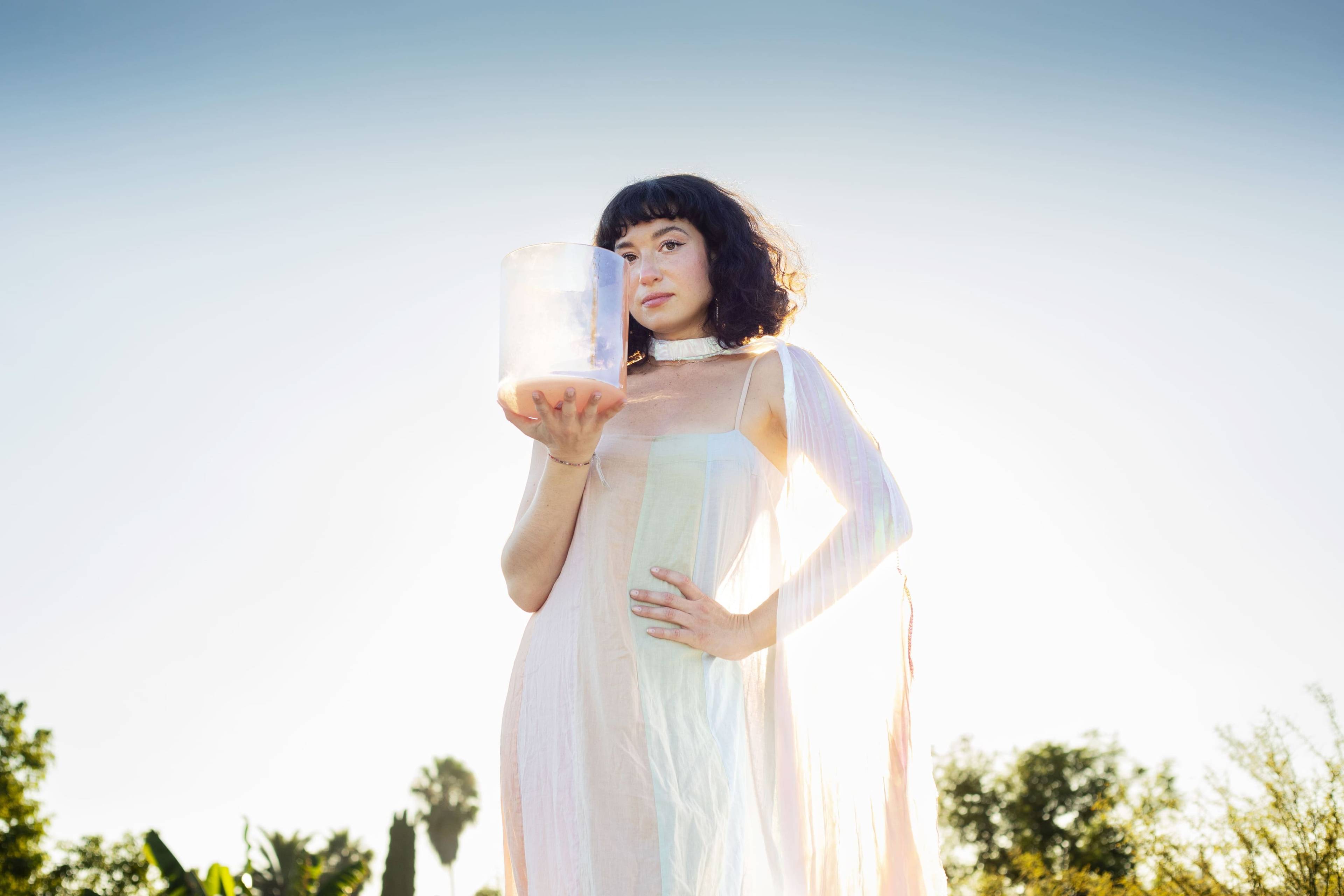 Woman in a light sheer dress holding a pale translucent cylindrical object outdoors.