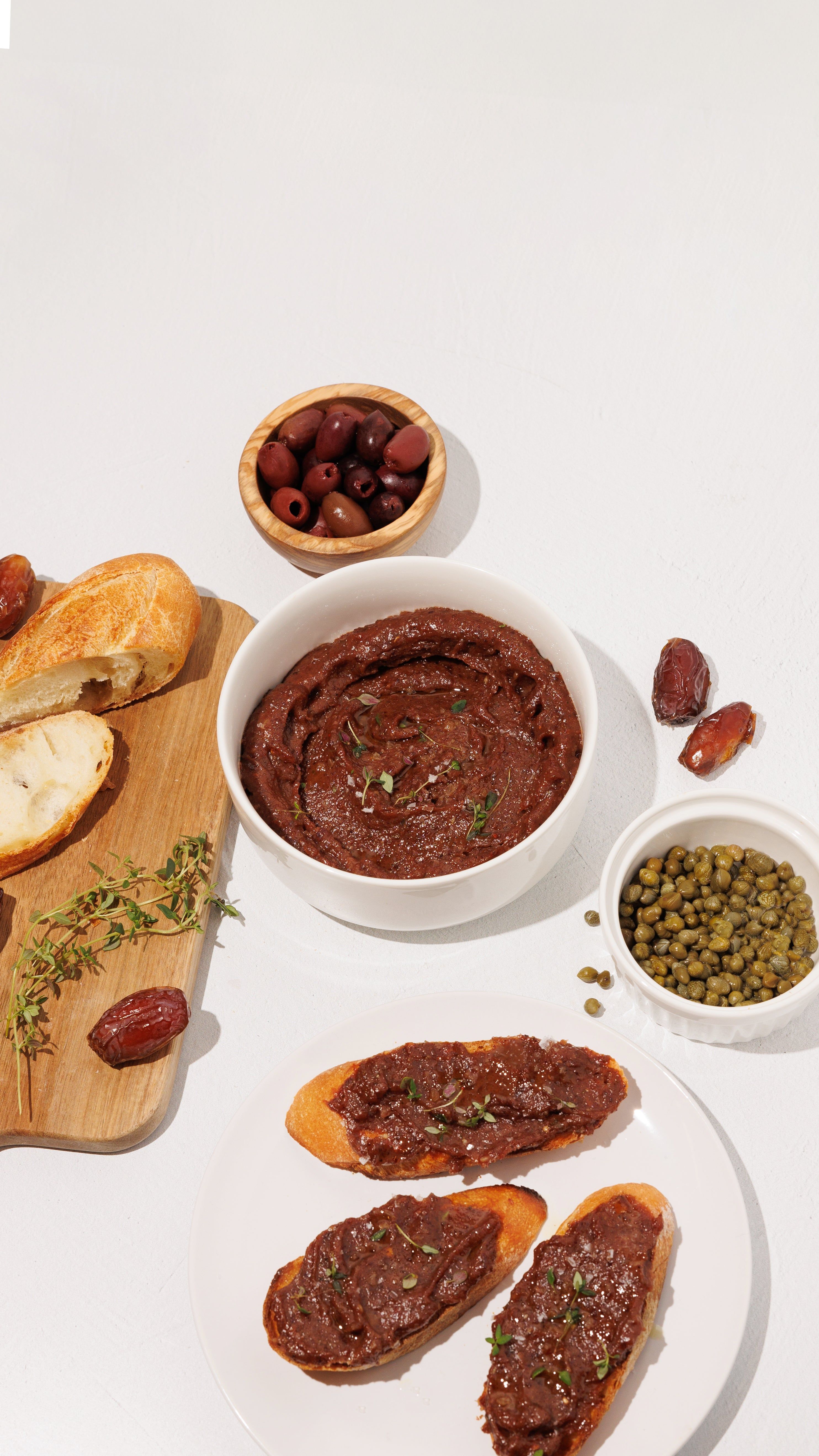 Bowls of olive date spread, olives, capers, dates, and toasted bread on a white table.