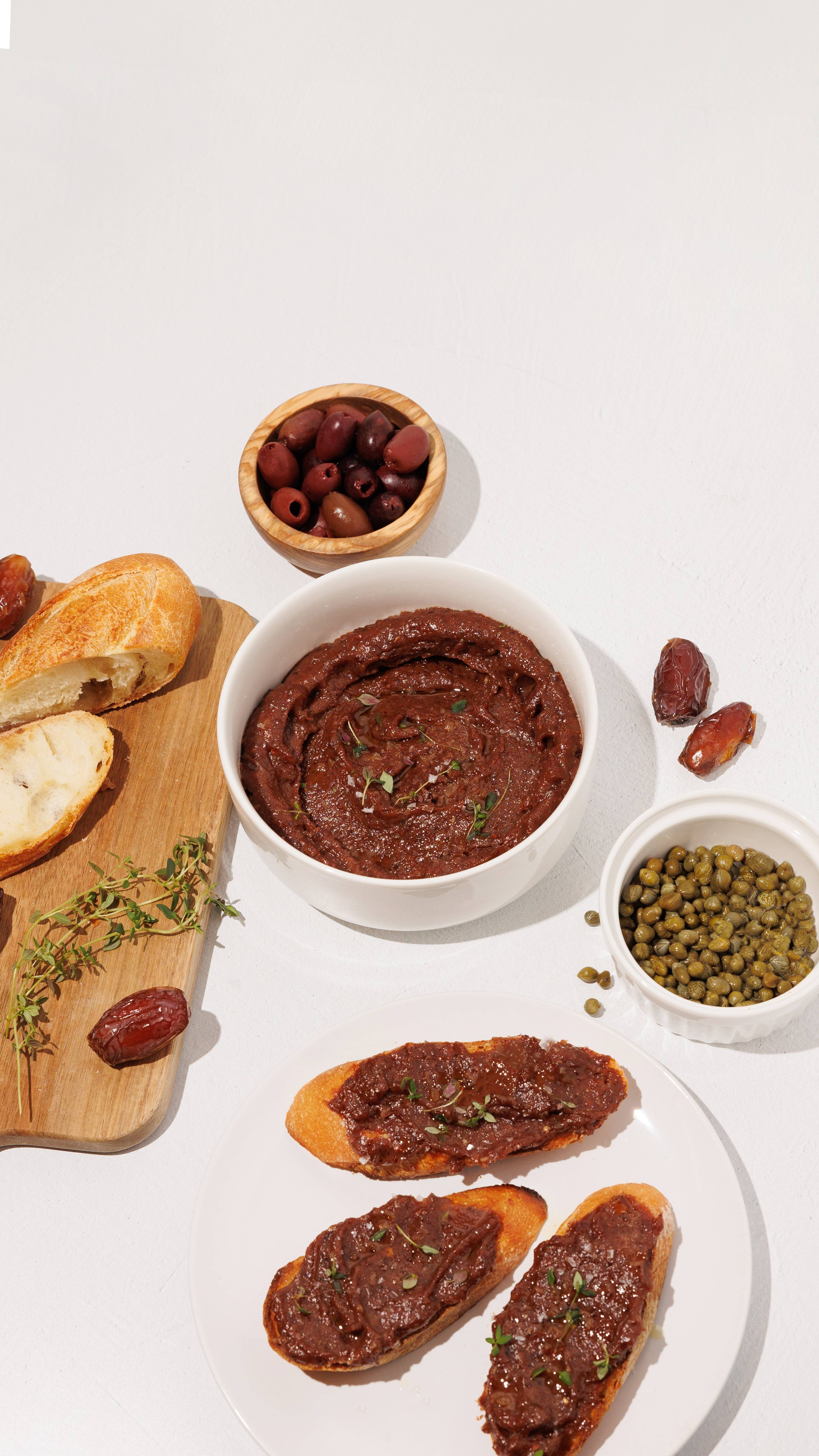 Bowls of olive date spread, olives, capers, dates, and toasted bread on a white table.
