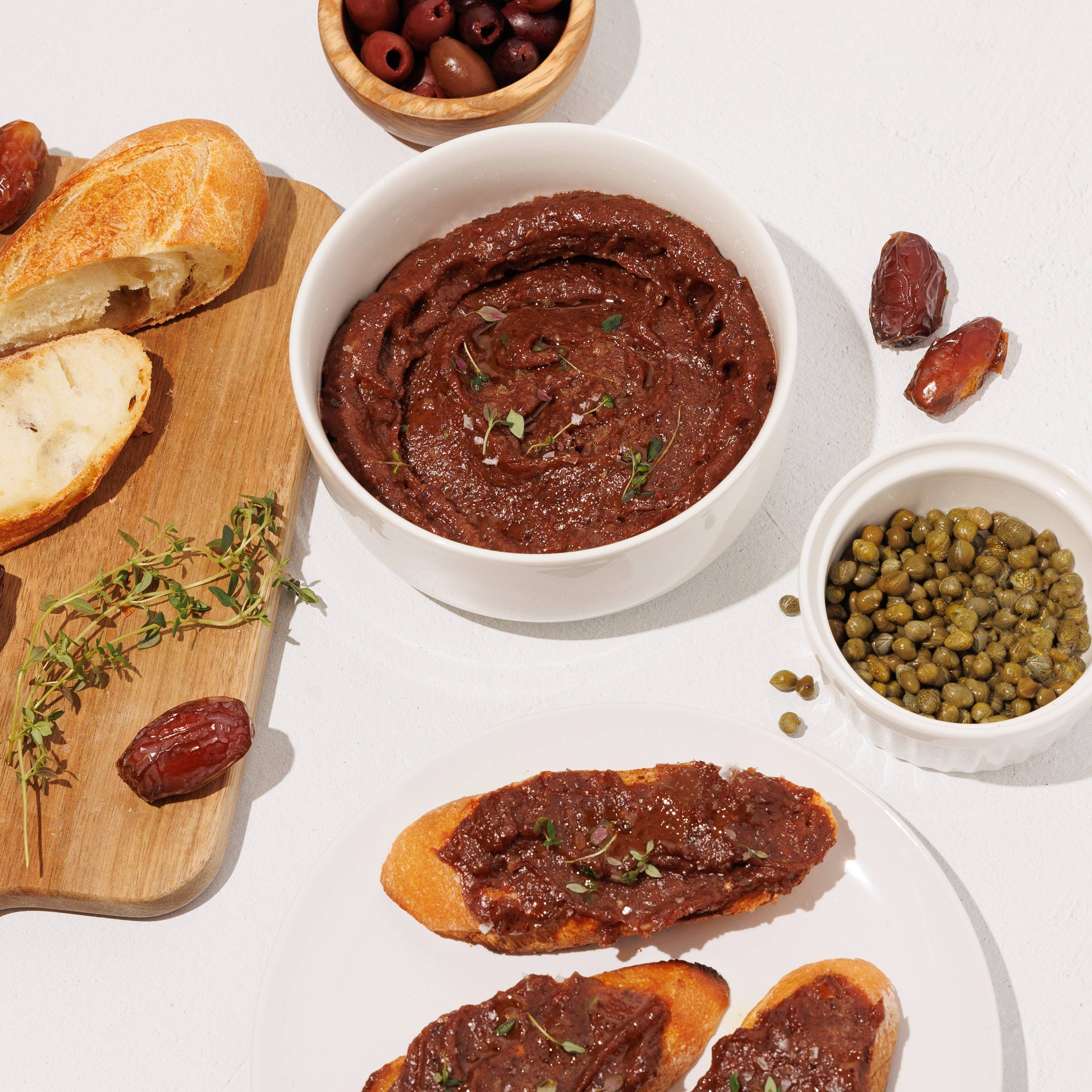 Bowls of olive date spread, olives, capers, dates, and toasted bread on a white table.