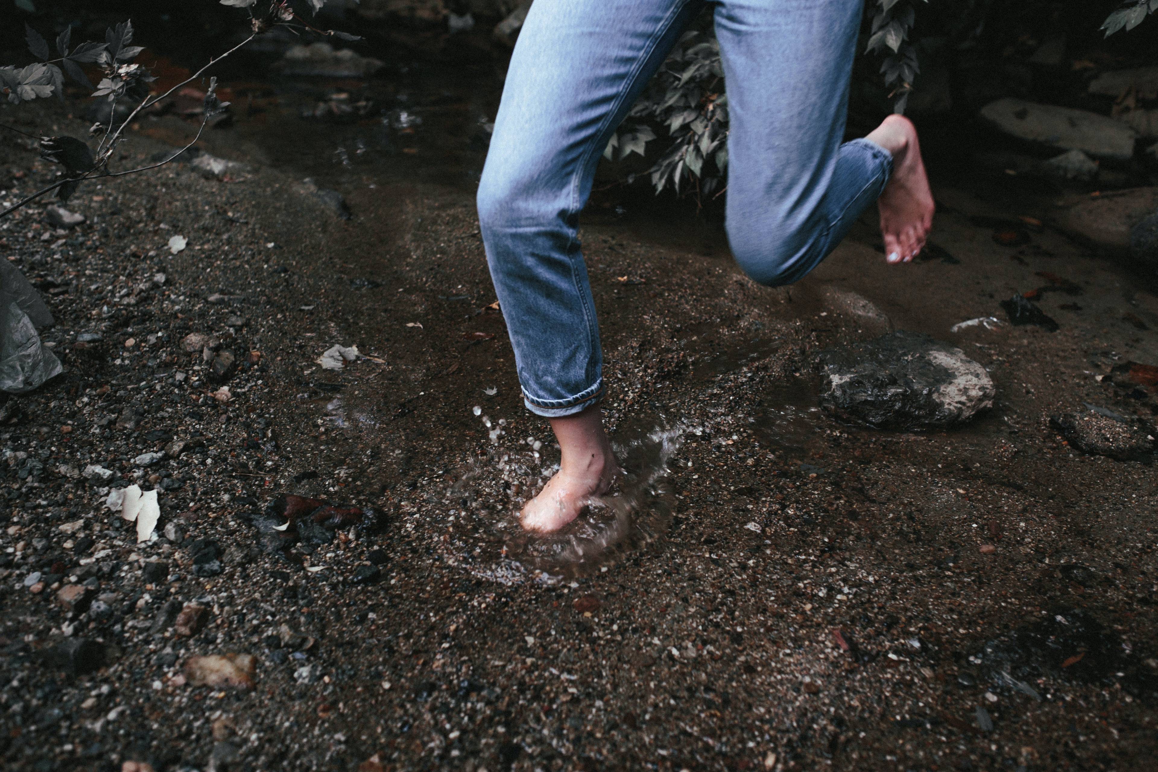 Bare feet in rolled-up jeans stepping through a shallow rocky stream, one foot raised.