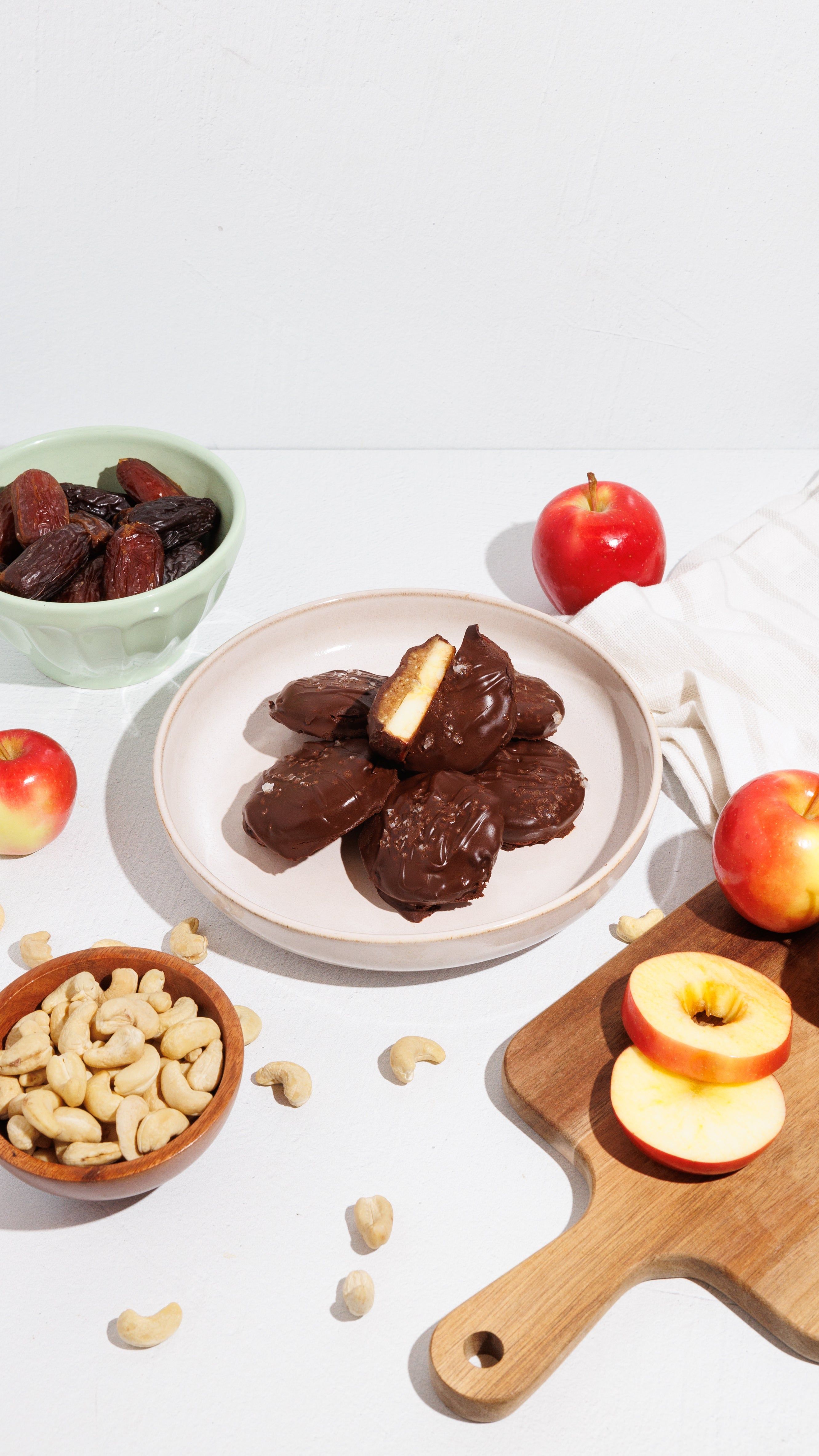 Chocolate-coated rounds on a plate with apples, dates, and cashews on a white tabletop.