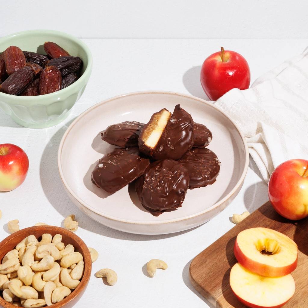 Chocolate-coated rounds on a plate with apples, dates, and cashews on a white tabletop.