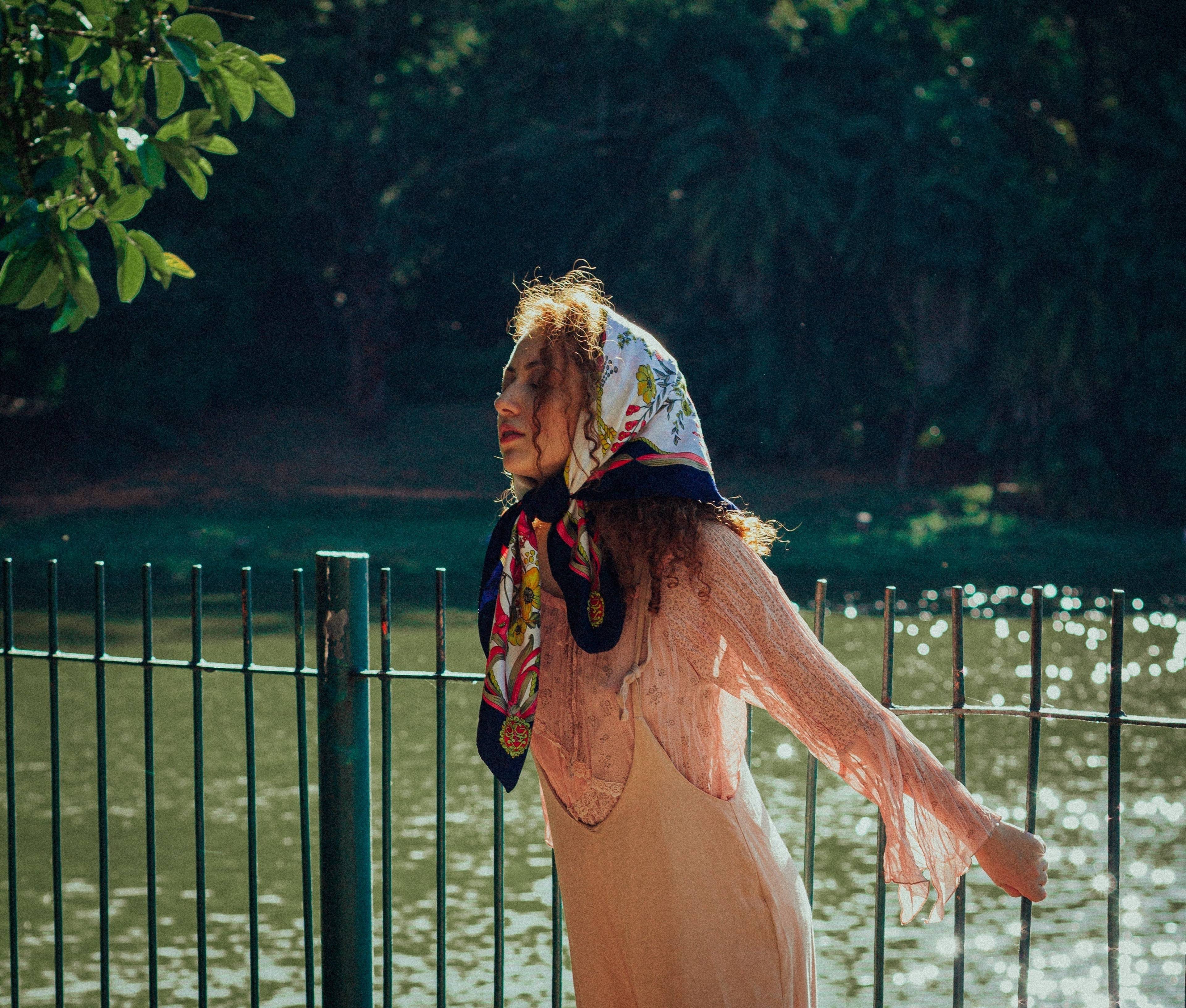 Woman in a patterned headscarf and peach sheer top over a tan dress stands beside a fence by sunlit water.