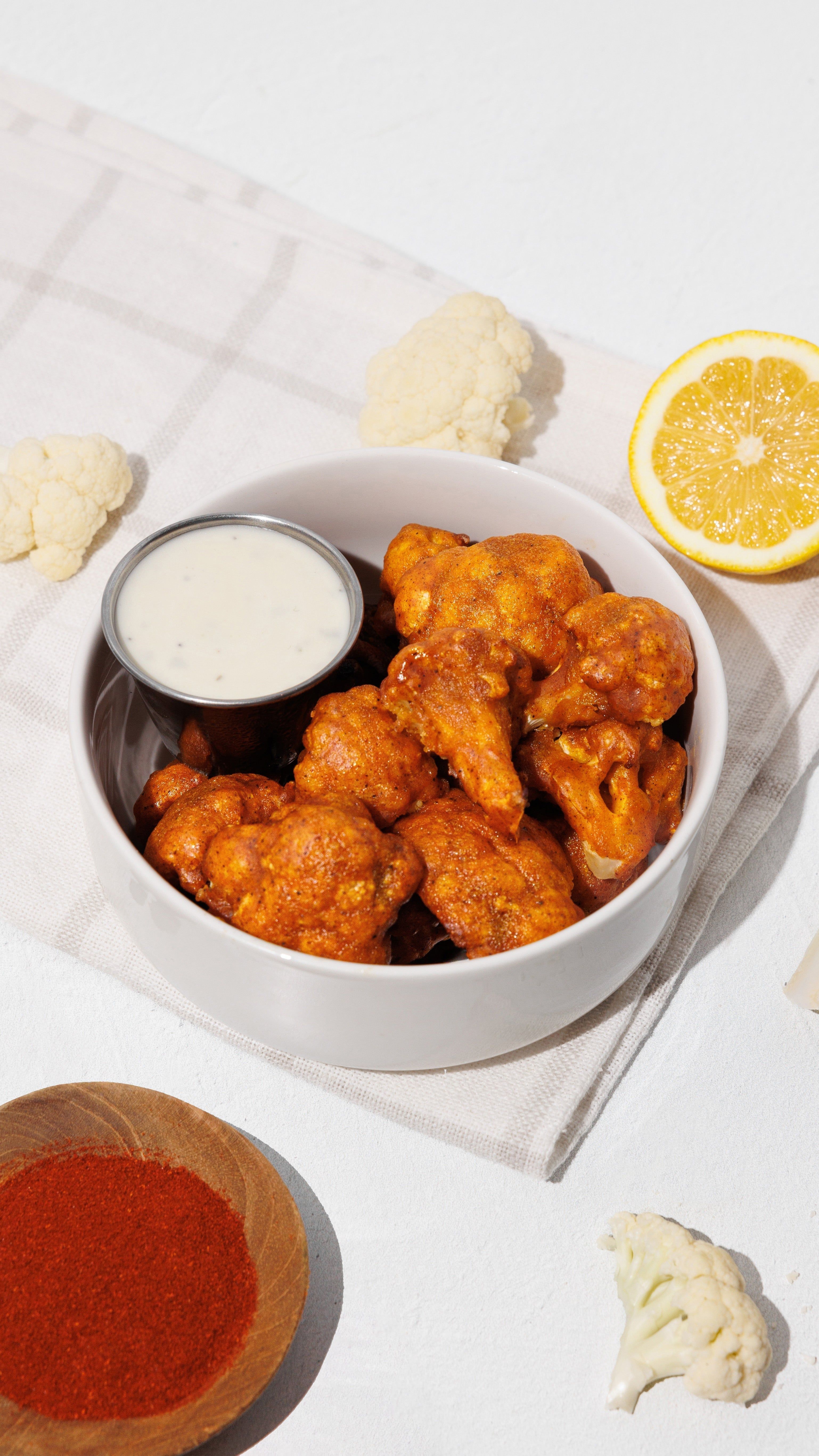 Bowl of buffalo cauliflower bites with creamy dipping sauce, lemon, cauliflower florets, and red seasoning.