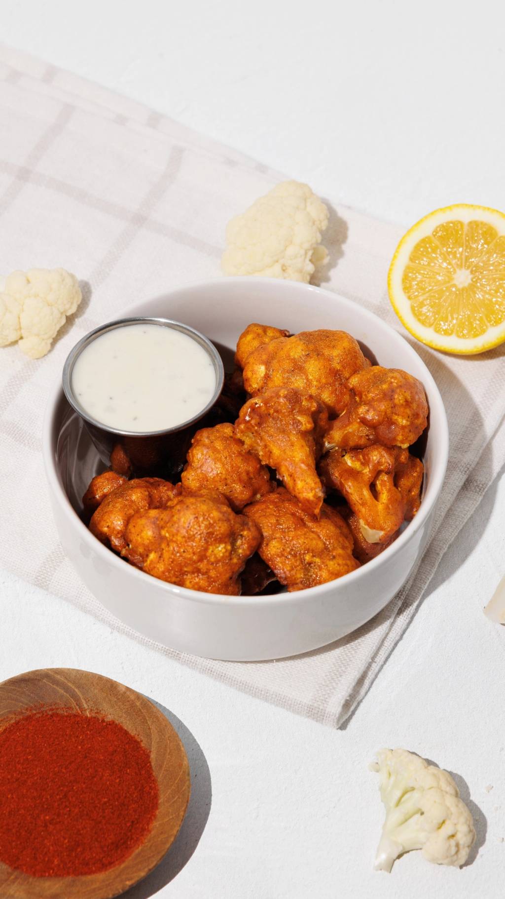 Bowl of buffalo cauliflower bites with creamy dipping sauce, lemon, cauliflower florets, and red seasoning.