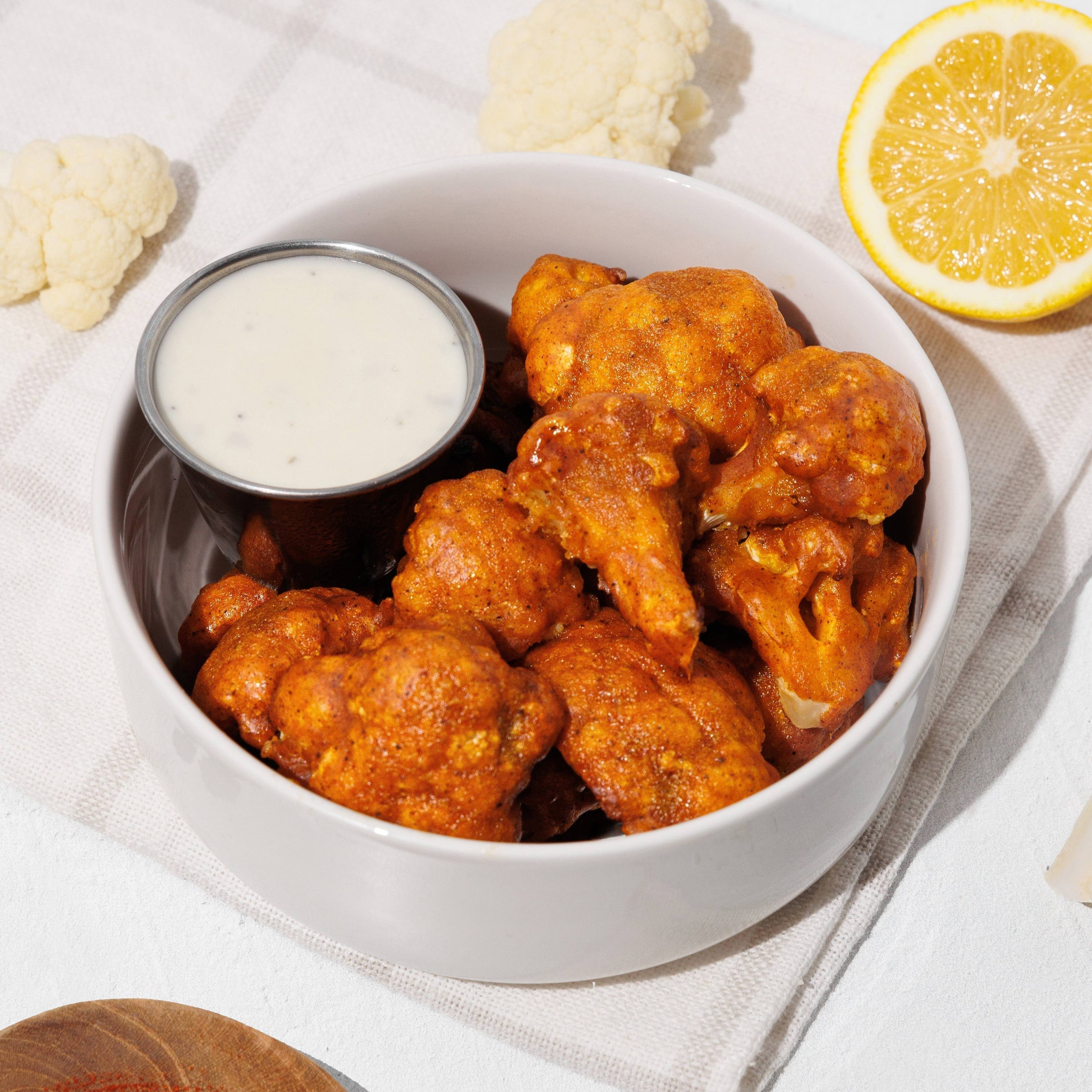 Bowl of buffalo cauliflower bites with creamy dipping sauce, lemon, cauliflower florets, and red seasoning.