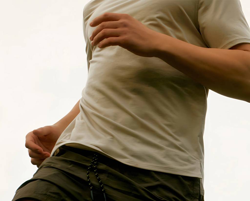Cropped person in a light-colored T-shirt and dark shorts, with one hand near the waist and the other across the chest.