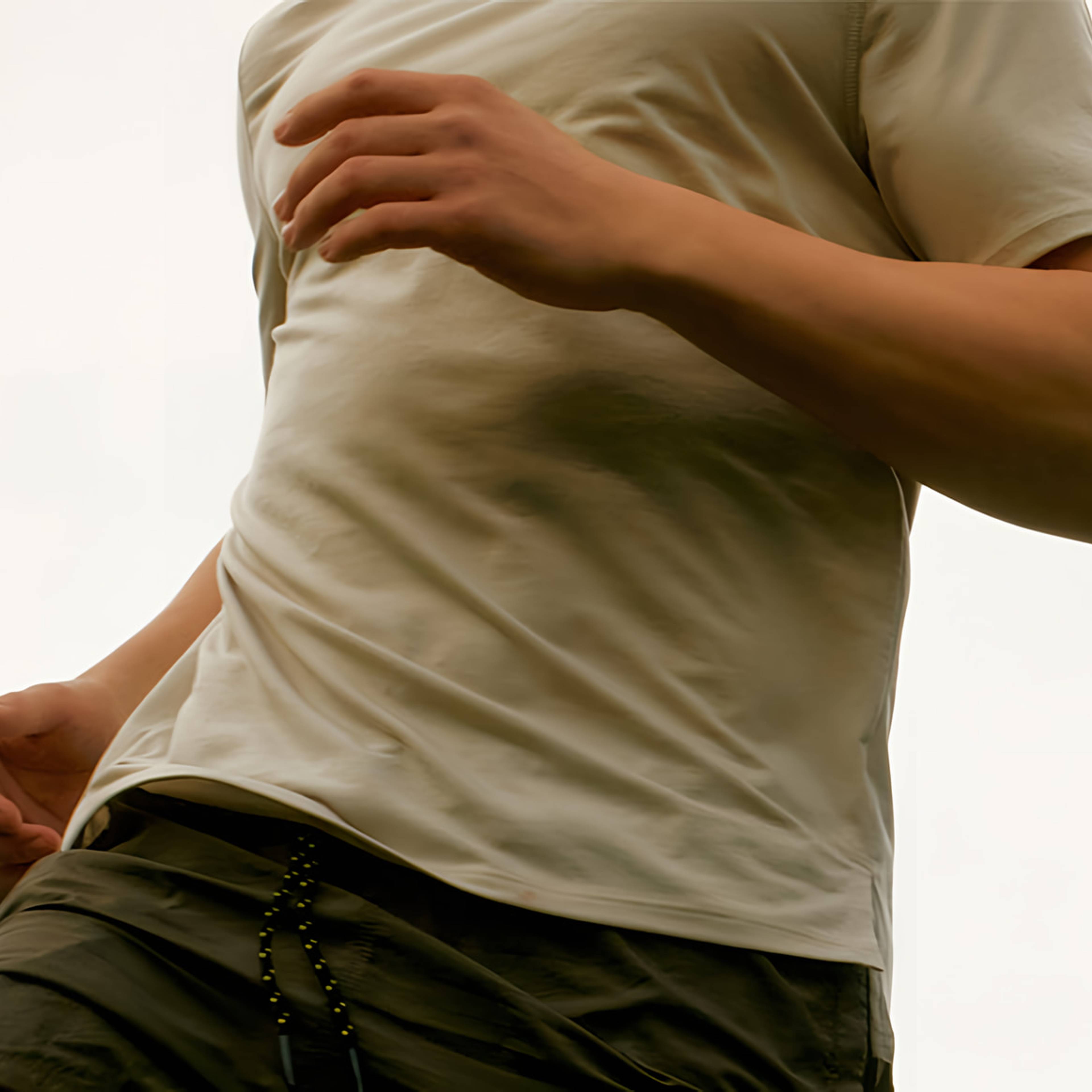 Cropped person in a light-colored T-shirt and dark shorts, with one hand near the waist and the other across the chest.