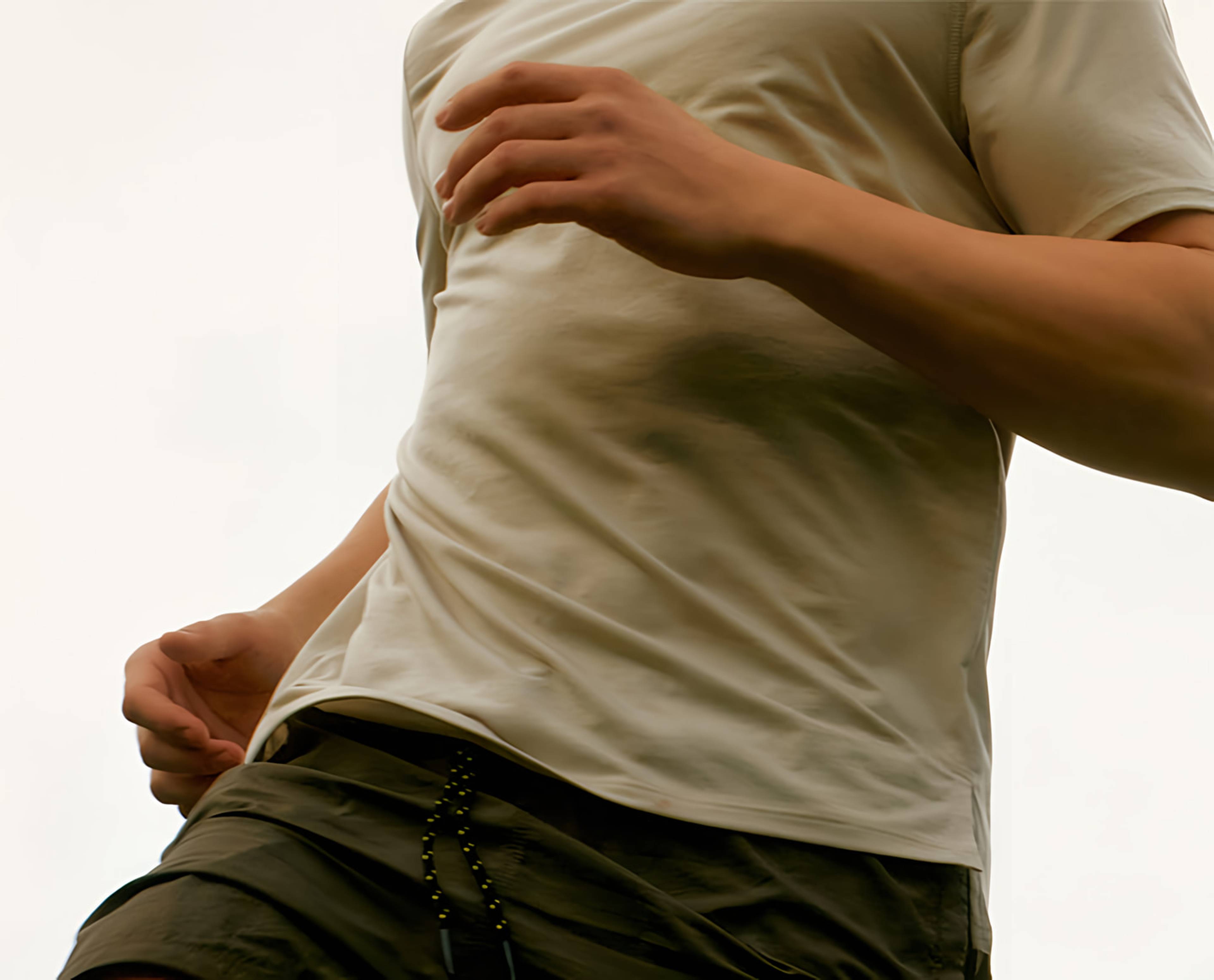Cropped person in a light-colored T-shirt and dark shorts, with one hand near the waist and the other across the chest.