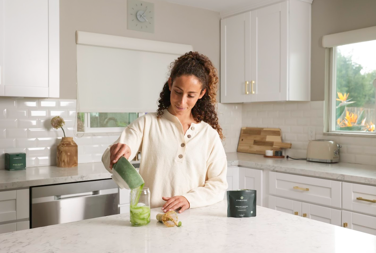 Woman pouring from a green container into a clear cup in a kitchen, with a dark pouch nearby.