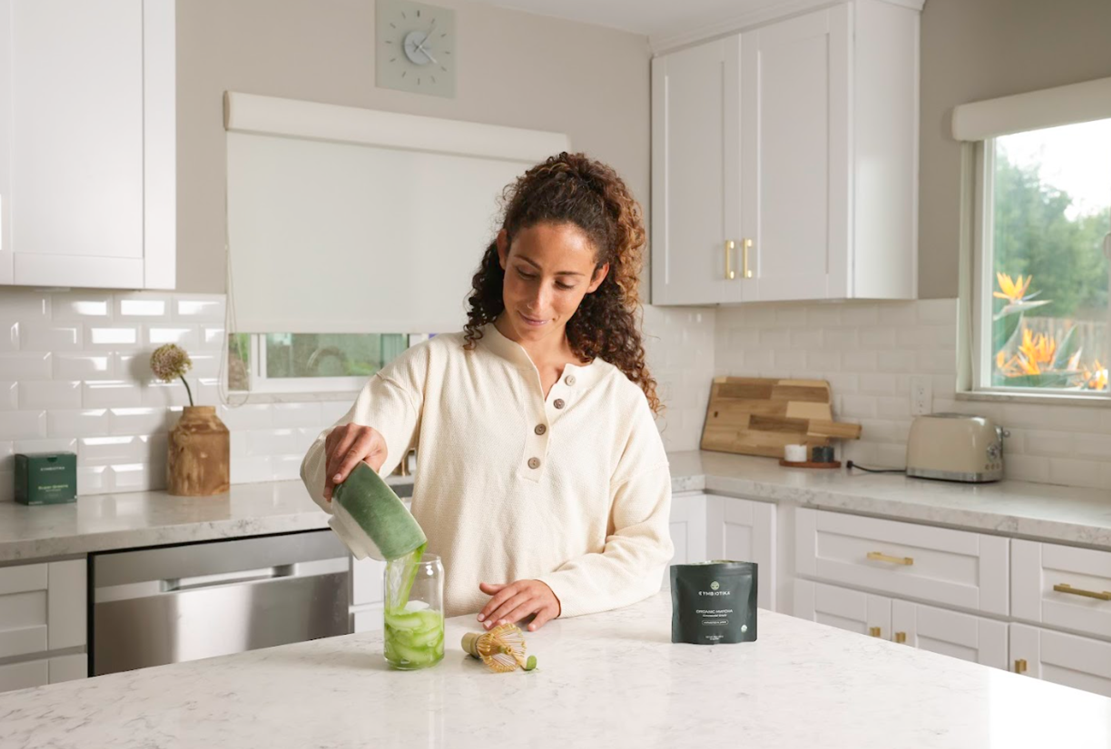 Woman pouring from a green container into a clear cup in a kitchen, with a dark pouch nearby.