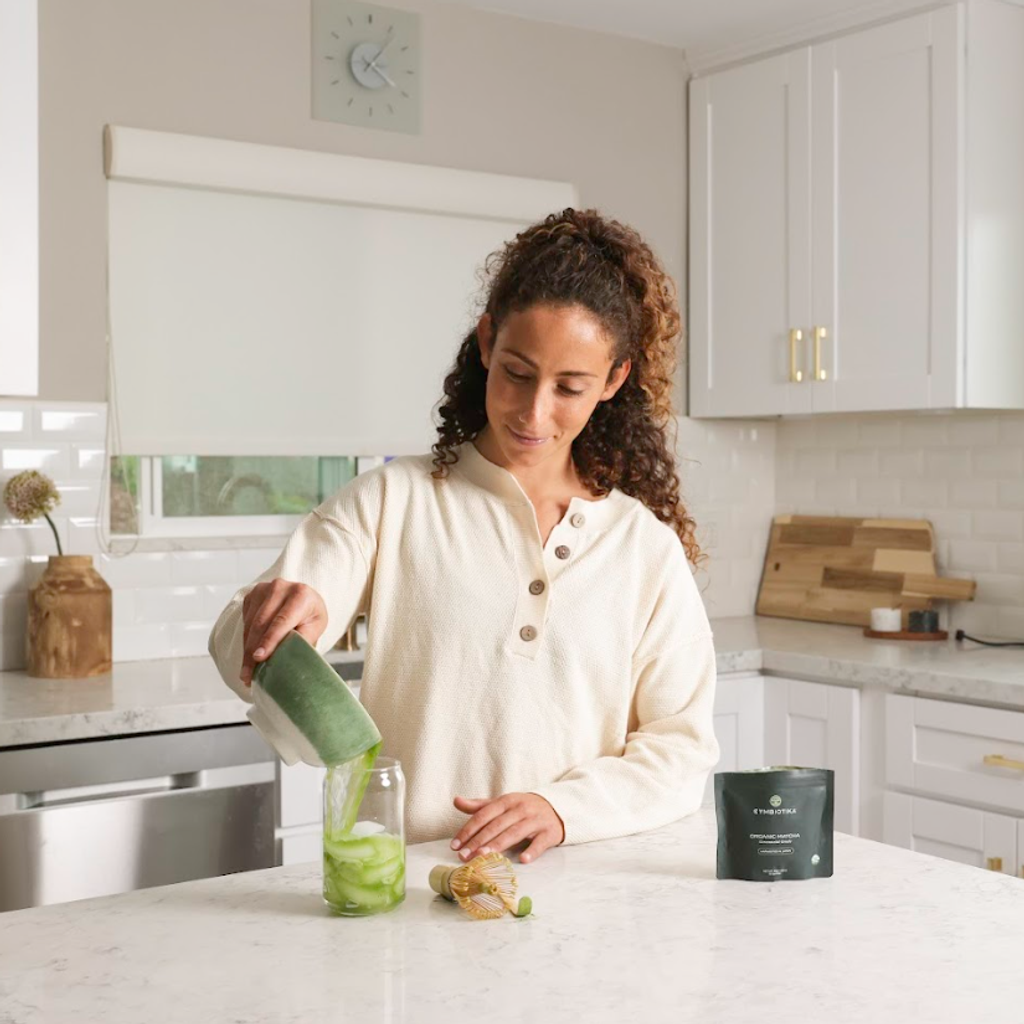 Woman pouring from a green container into a clear cup in a kitchen, with a dark pouch nearby.