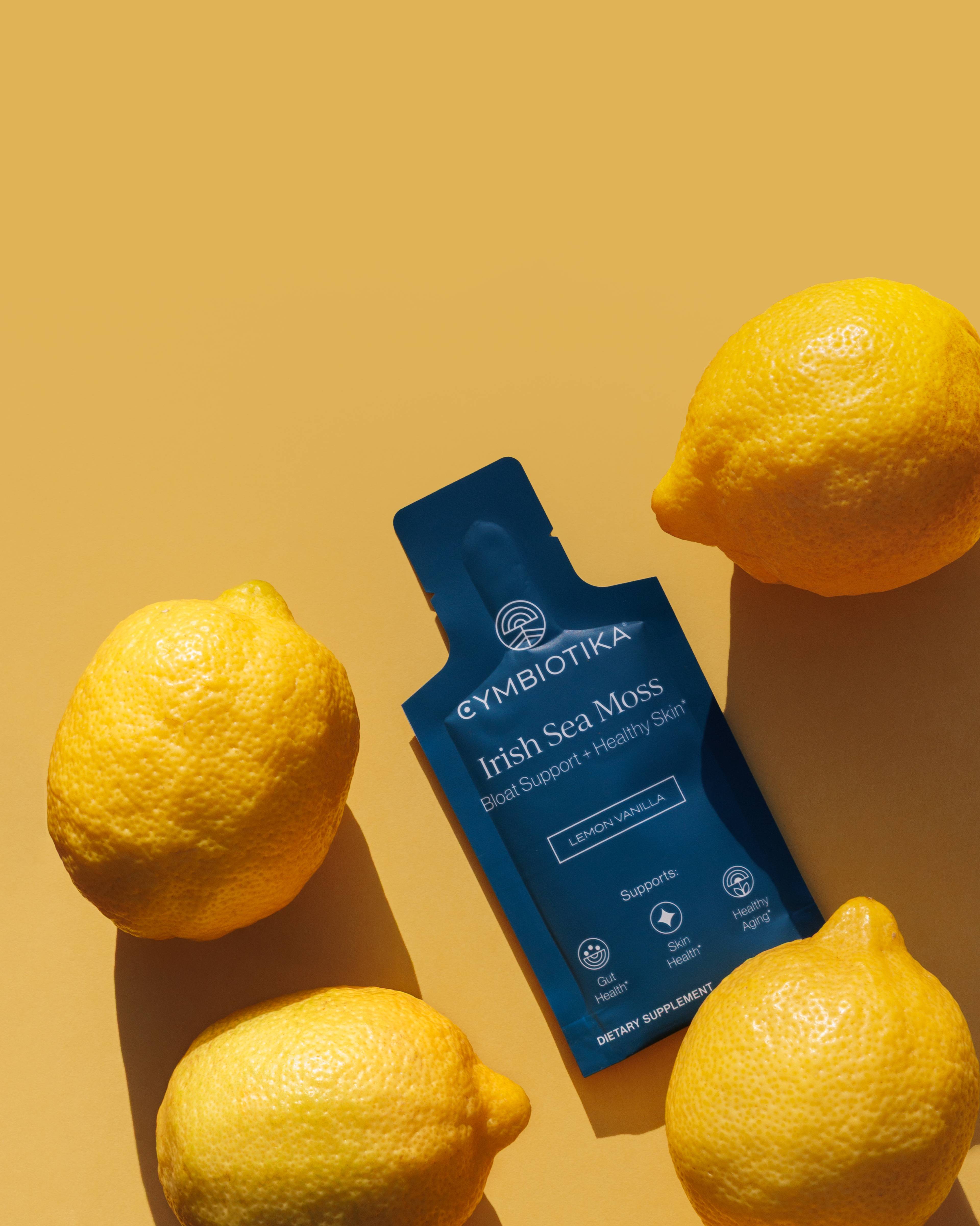 Blue Cymbiotika Irish Sea Moss single-serve packet labeled Lemon Vanilla, surrounded by lemons on a yellow background.