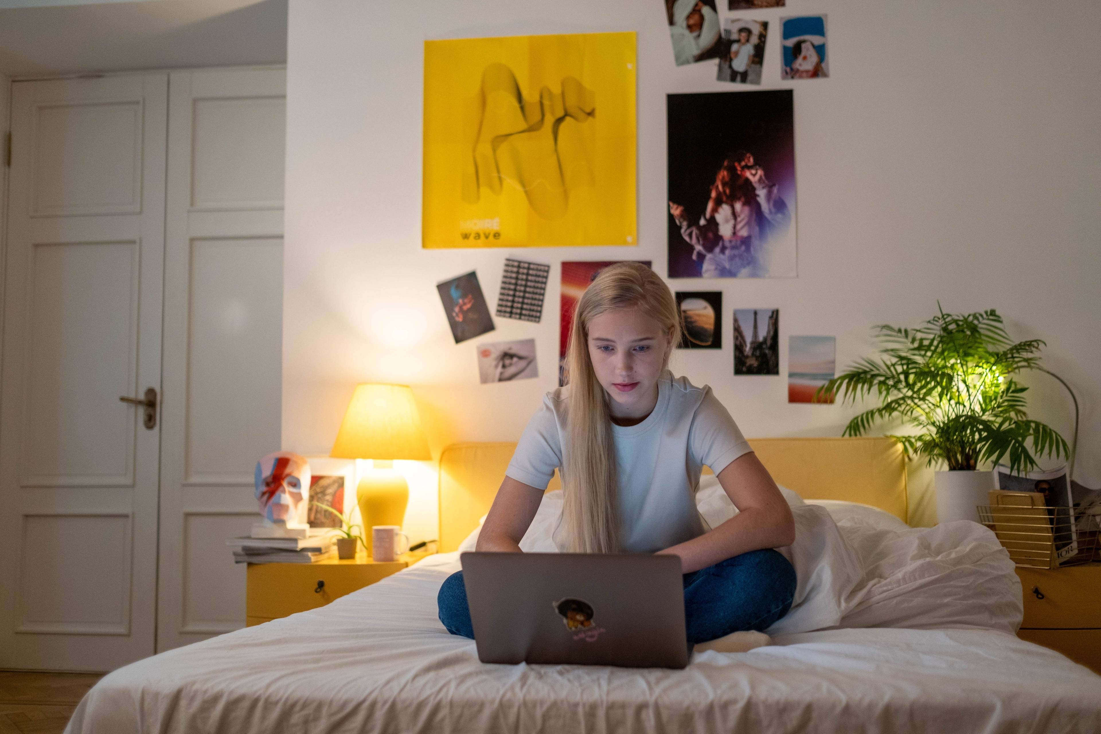 Young woman sitting on a bed and using a laptop in a softly lit bedroom.