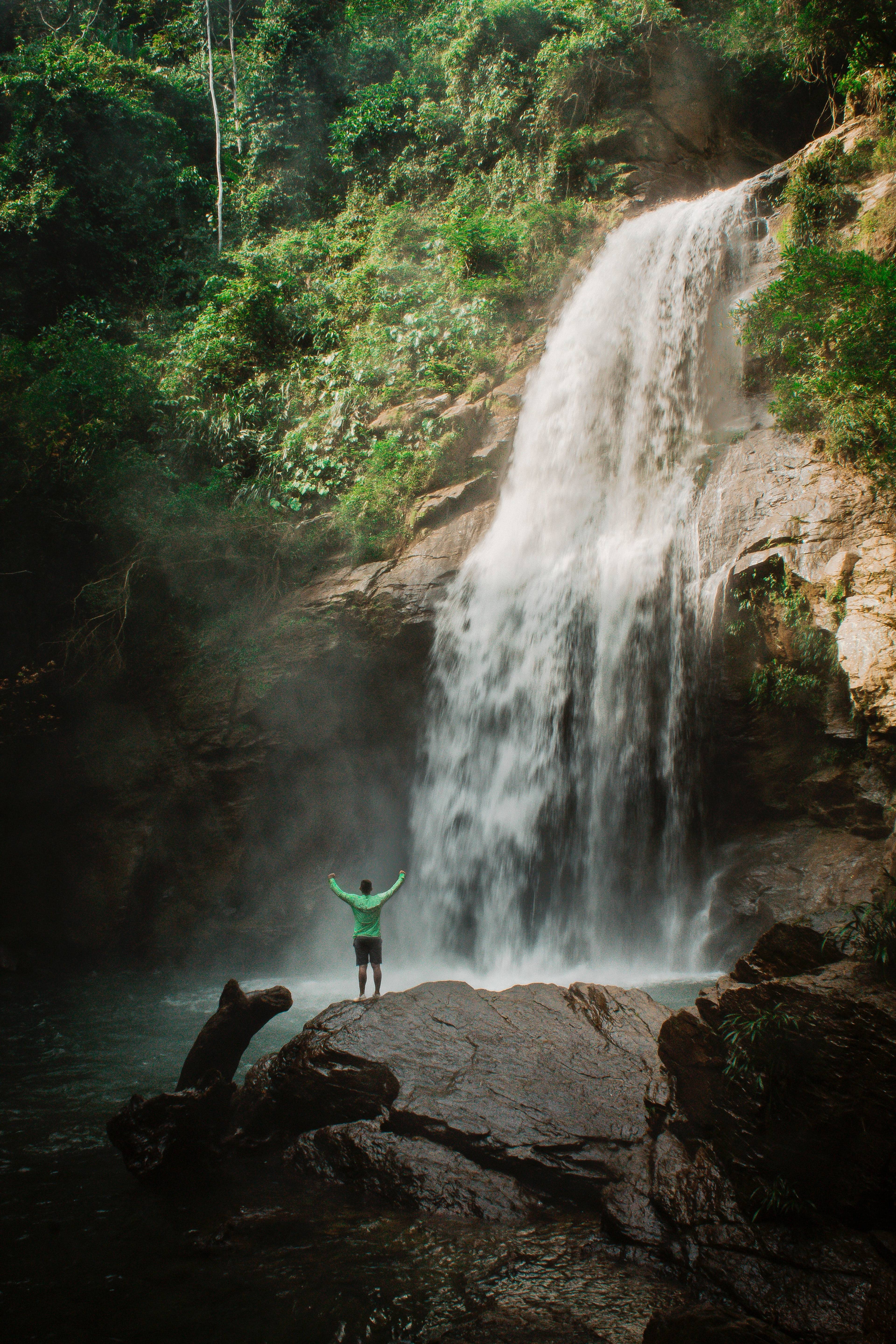 Person in a green top stands on a rock with arms raised before a forest waterfall.