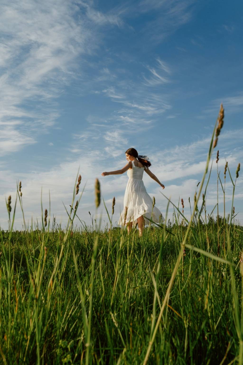 Woman in a white dress walking through a green field beneath a wide blue sky.