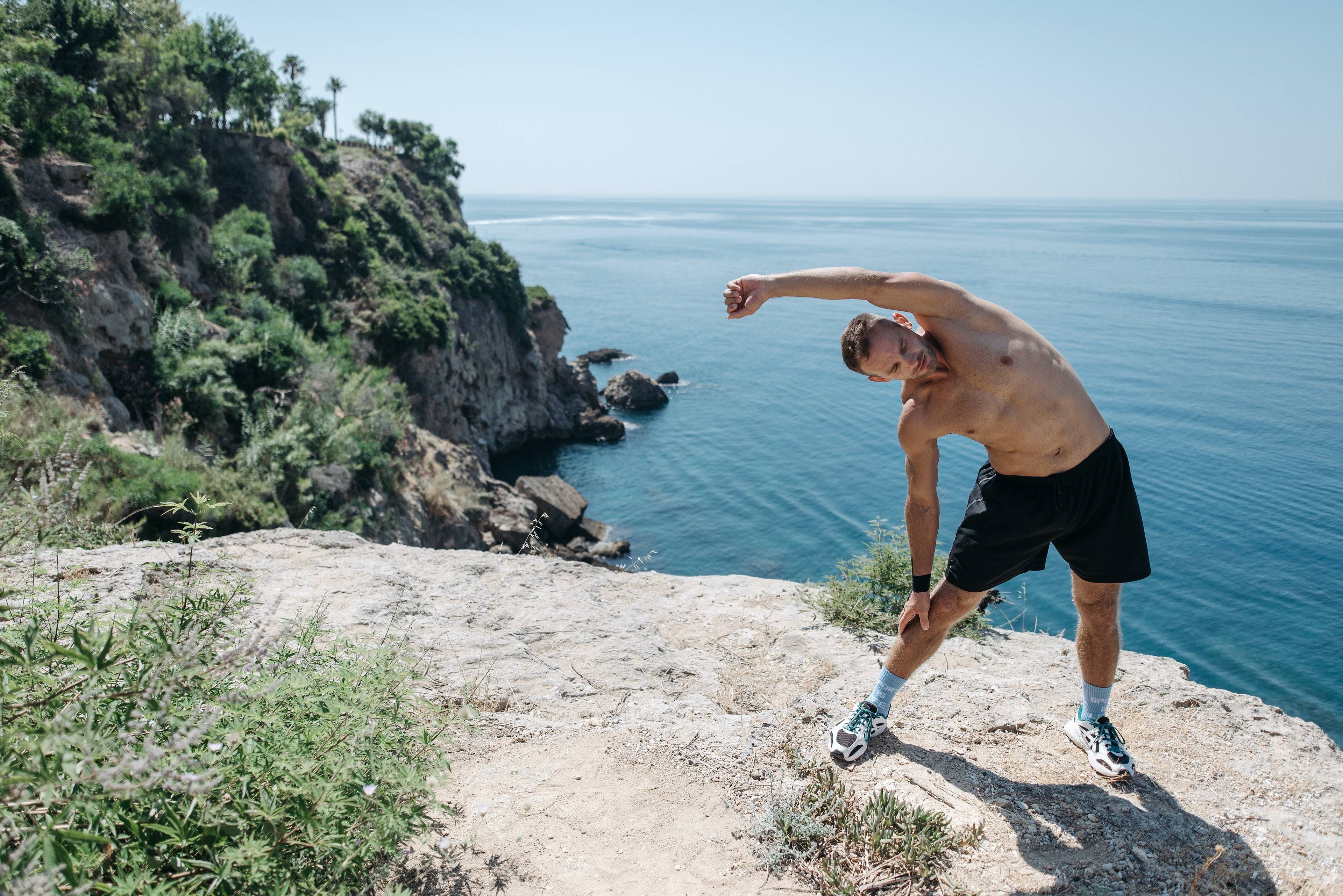 Shirtless man stretching on a rocky cliff above the ocean.