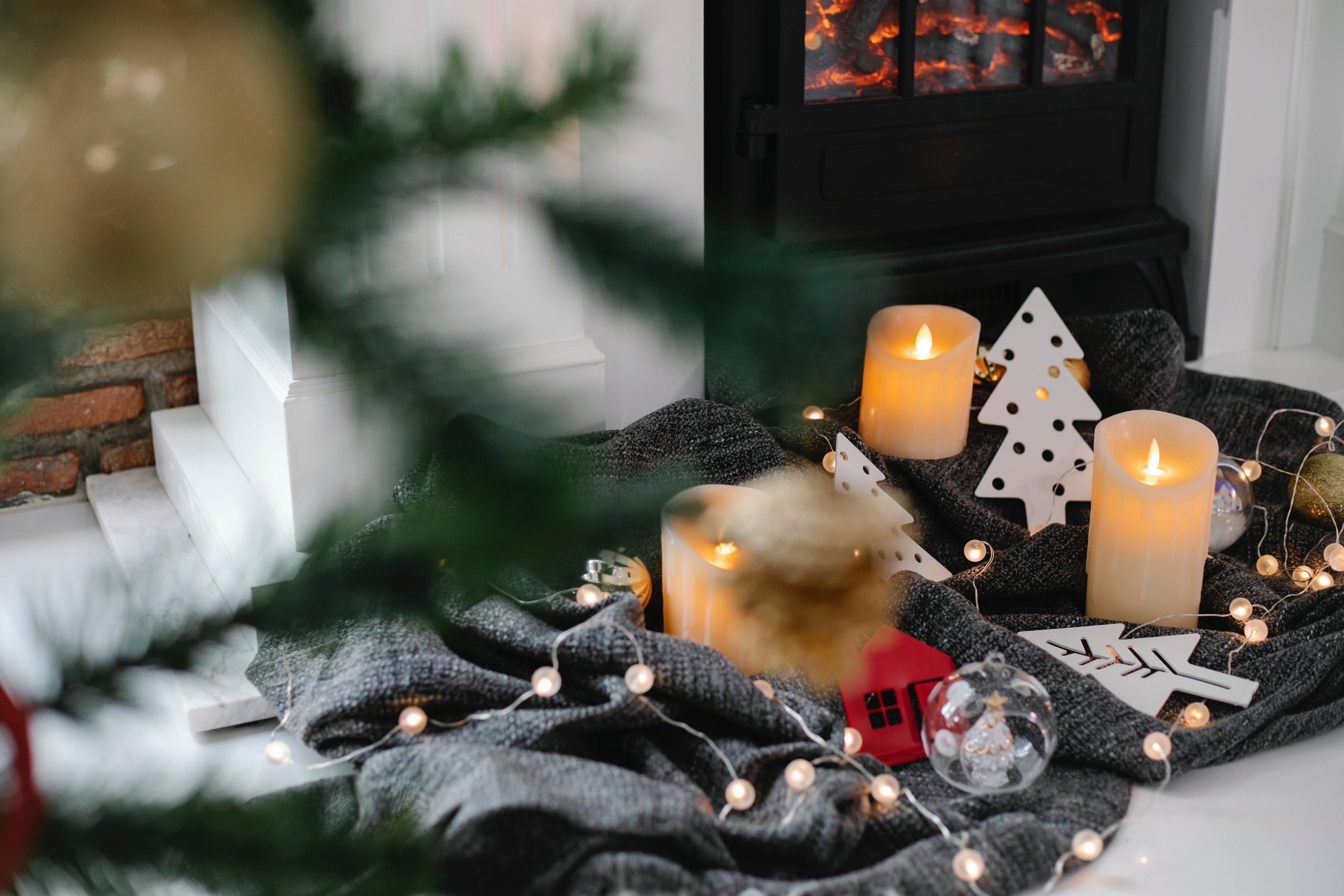 Lit candles, paper tree decorations, string lights, and a gray blanket beside a glowing fireplace.