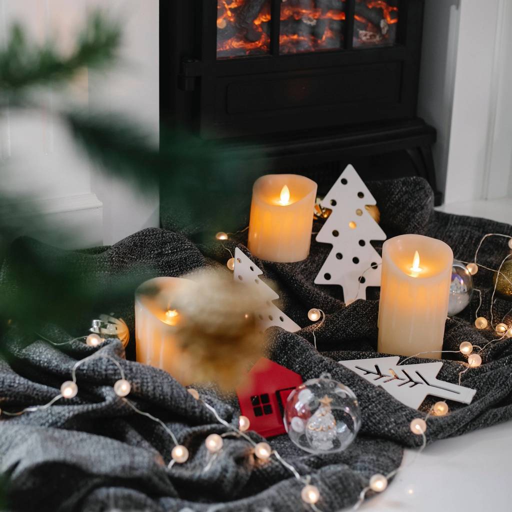 Lit candles, paper tree decorations, string lights, and a gray blanket beside a glowing fireplace.