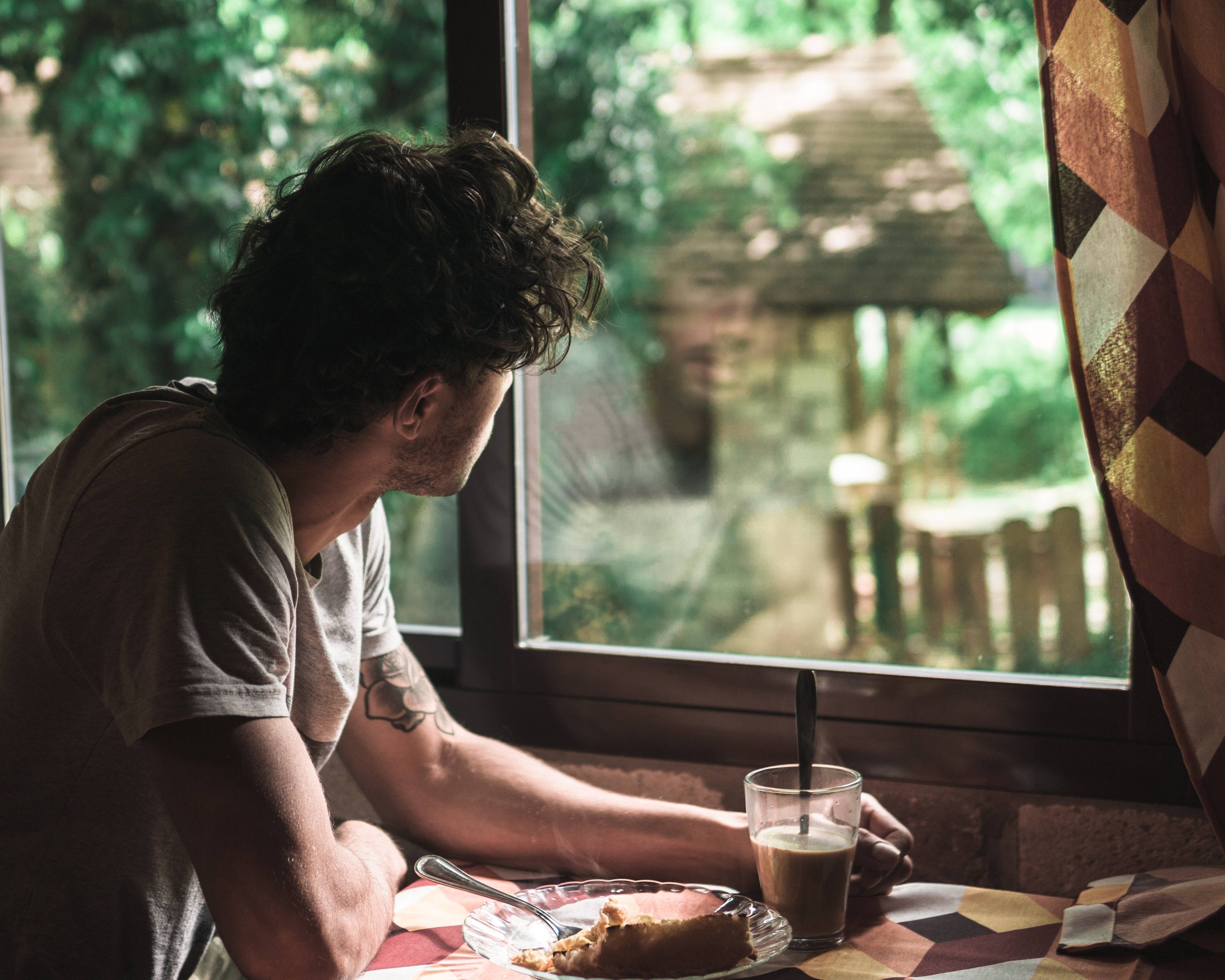 A man sits by a window with a mug and plate of food, looking out at a green garden.