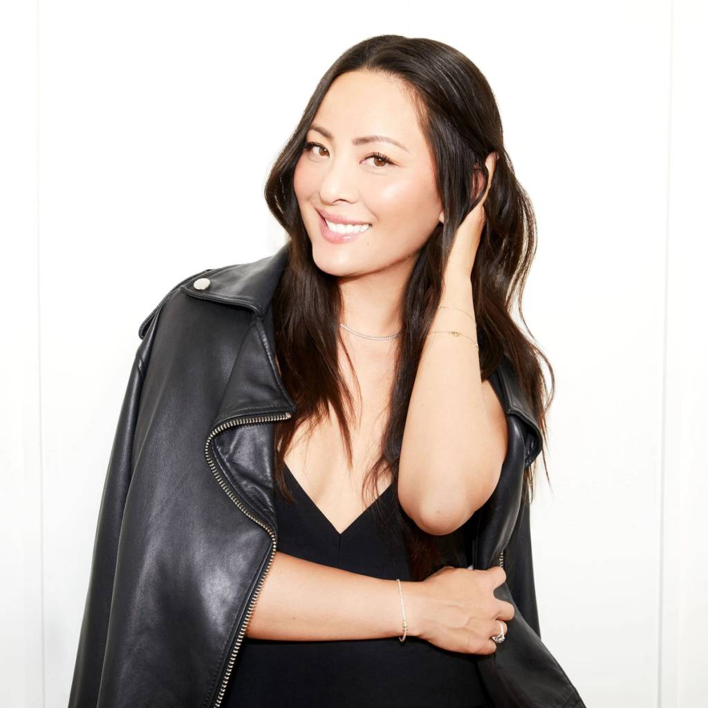 Smiling woman in a black dress with a leather jacket draped over her shoulders against a white wall.