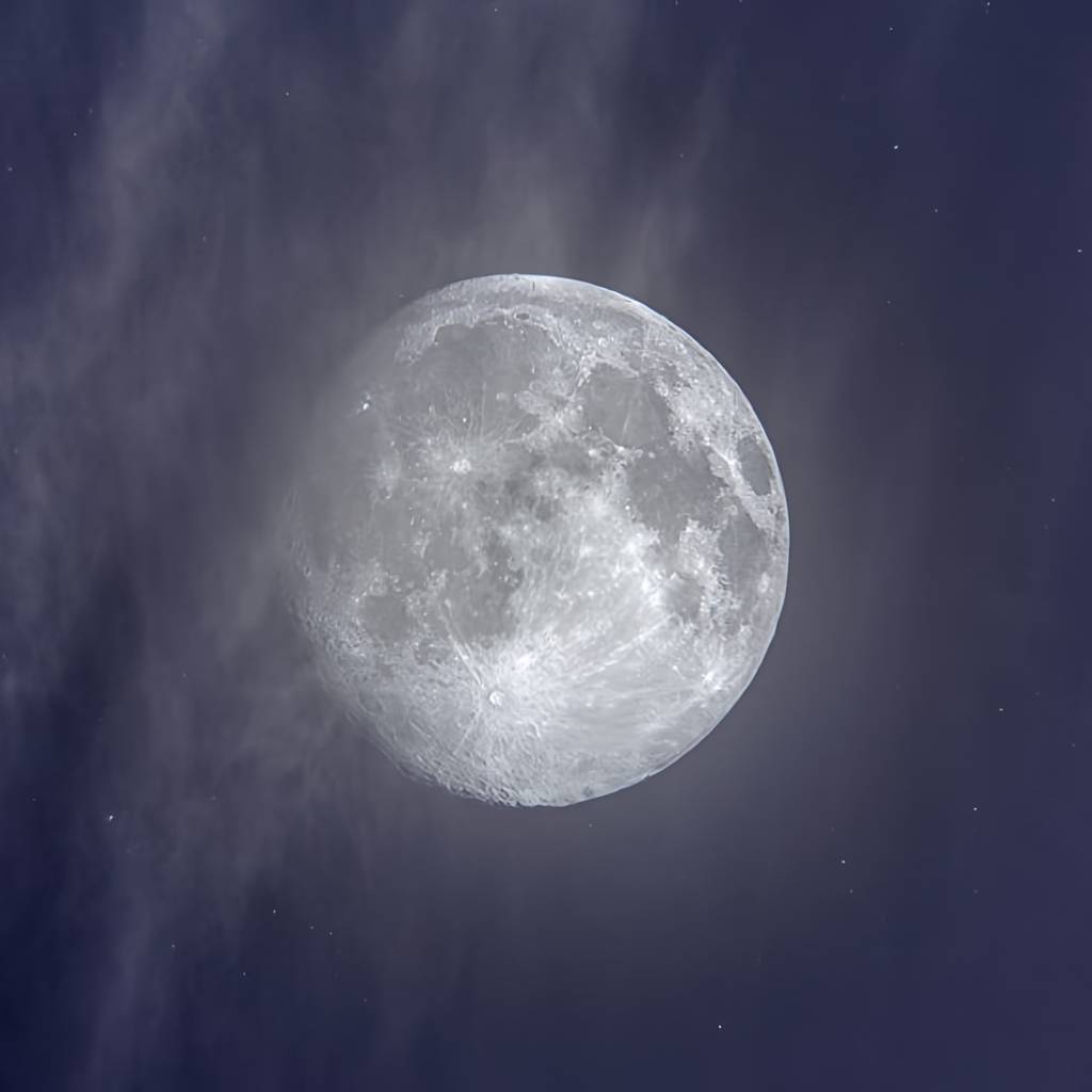 Bright full moon amid wispy clouds in a dark blue night sky.