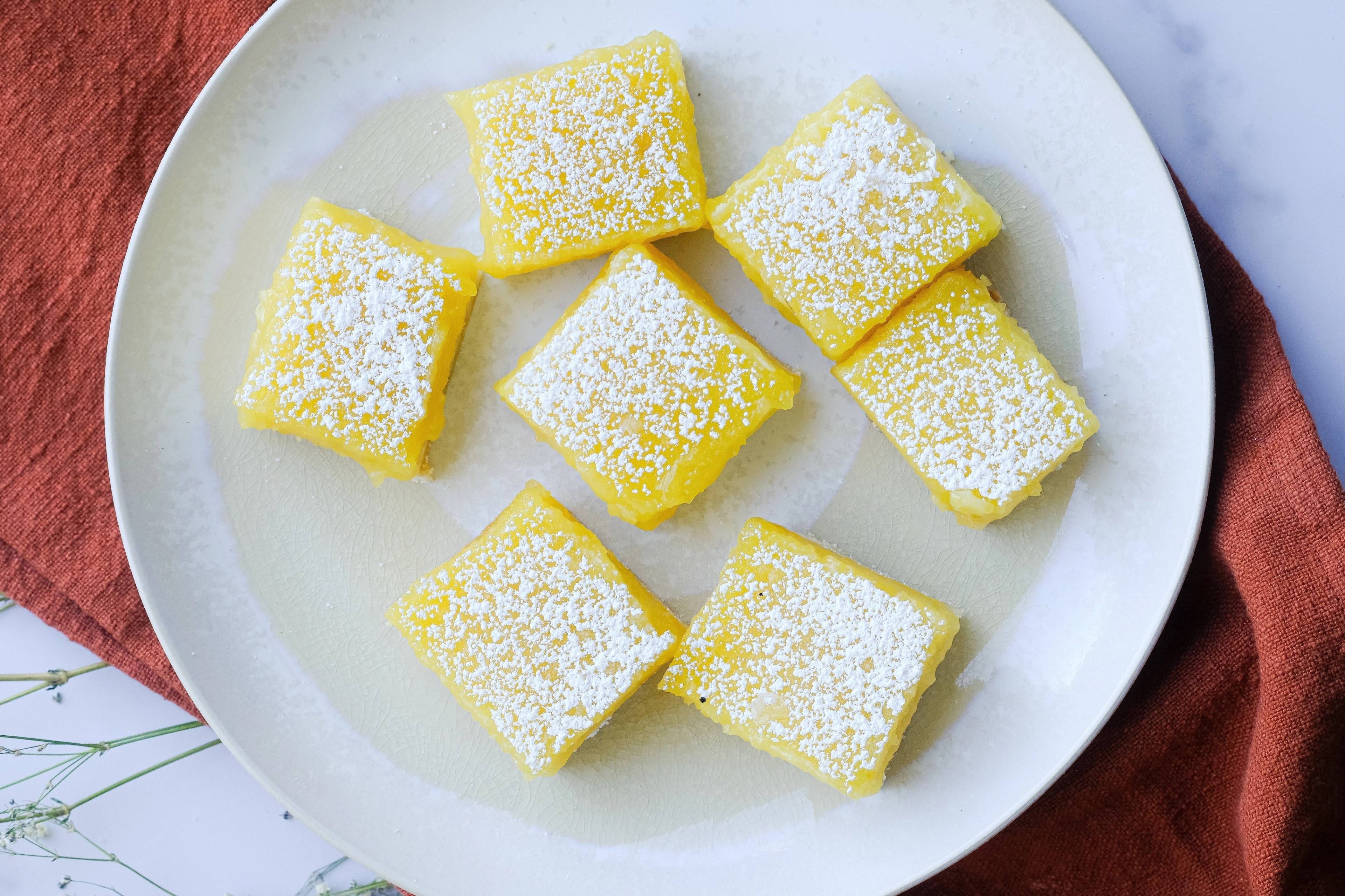 Seven powdered sugar-dusted lemon bars on a white plate with rust-colored fabric beneath it.