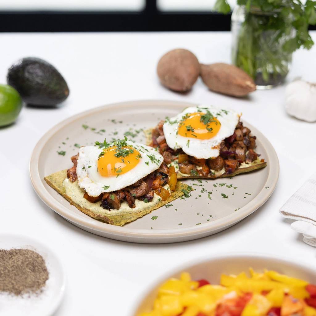 Two breakfast flatbreads topped with sautéed vegetables, fried eggs, and chopped herbs on a plate.