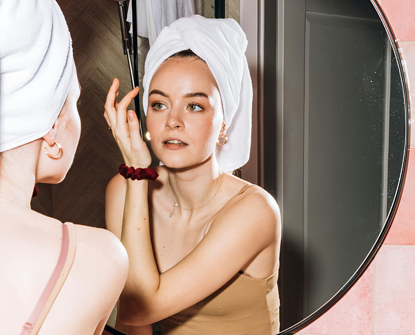 Woman with a towel wrapped around her hair looks at her reflection with one hand raised beside her face.