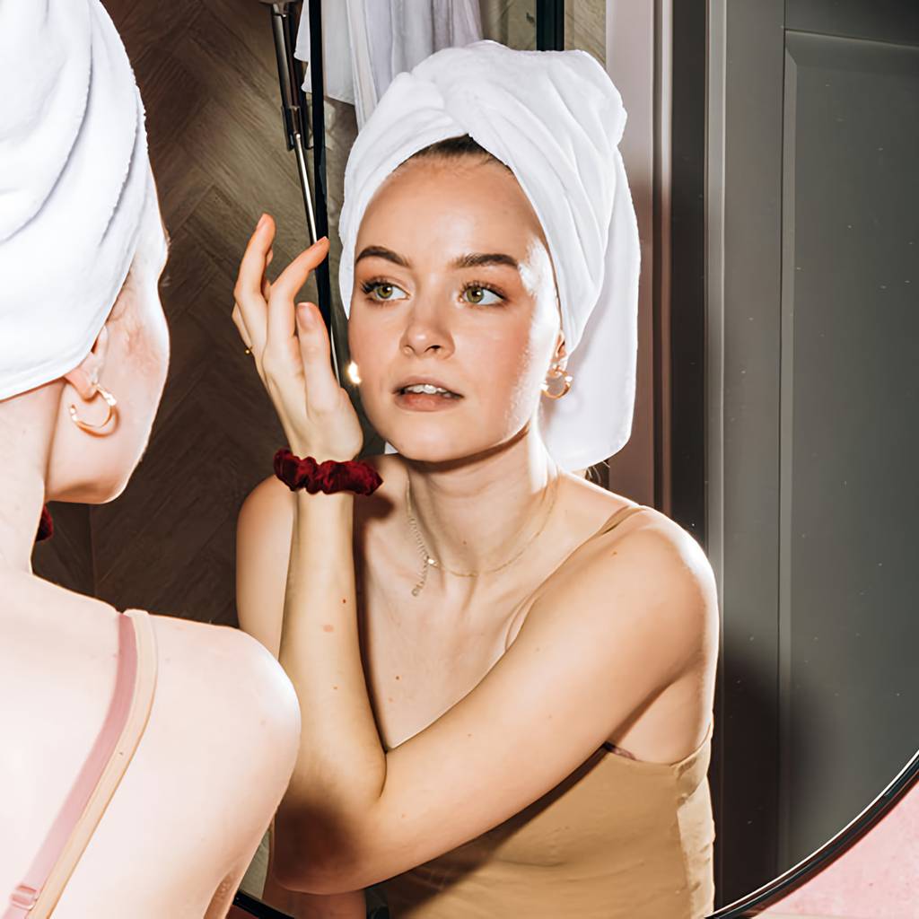 Woman with a towel wrapped around her hair looks at her reflection with one hand raised beside her face.