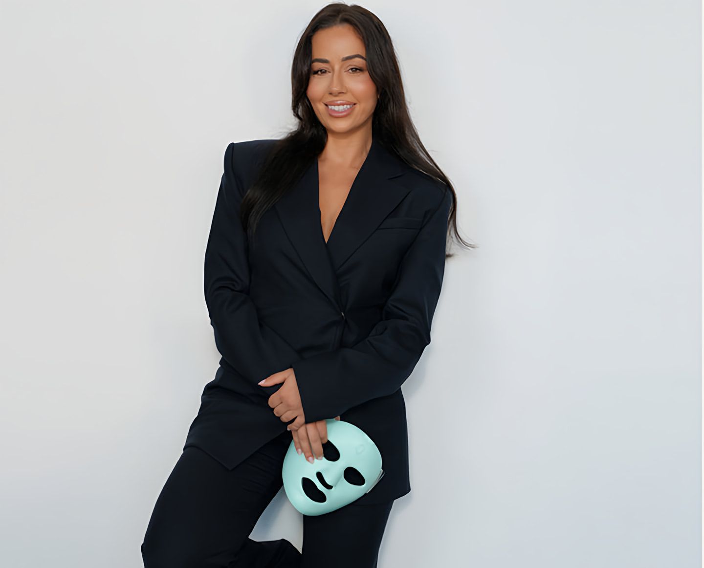 Smiling woman in a black suit holding a mint-green facial device against a white wall.