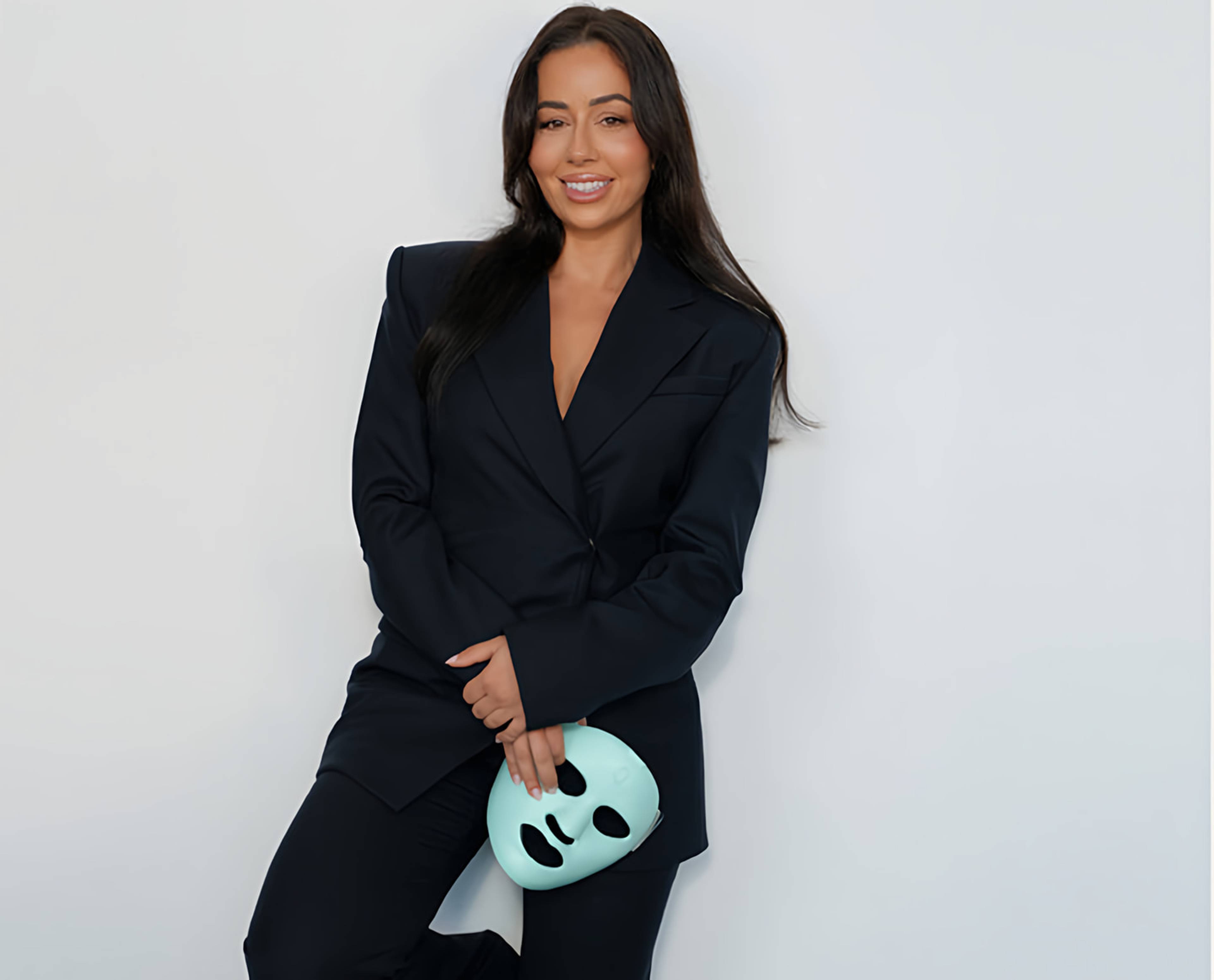 Smiling woman in a black suit holding a mint-green facial device against a white wall.