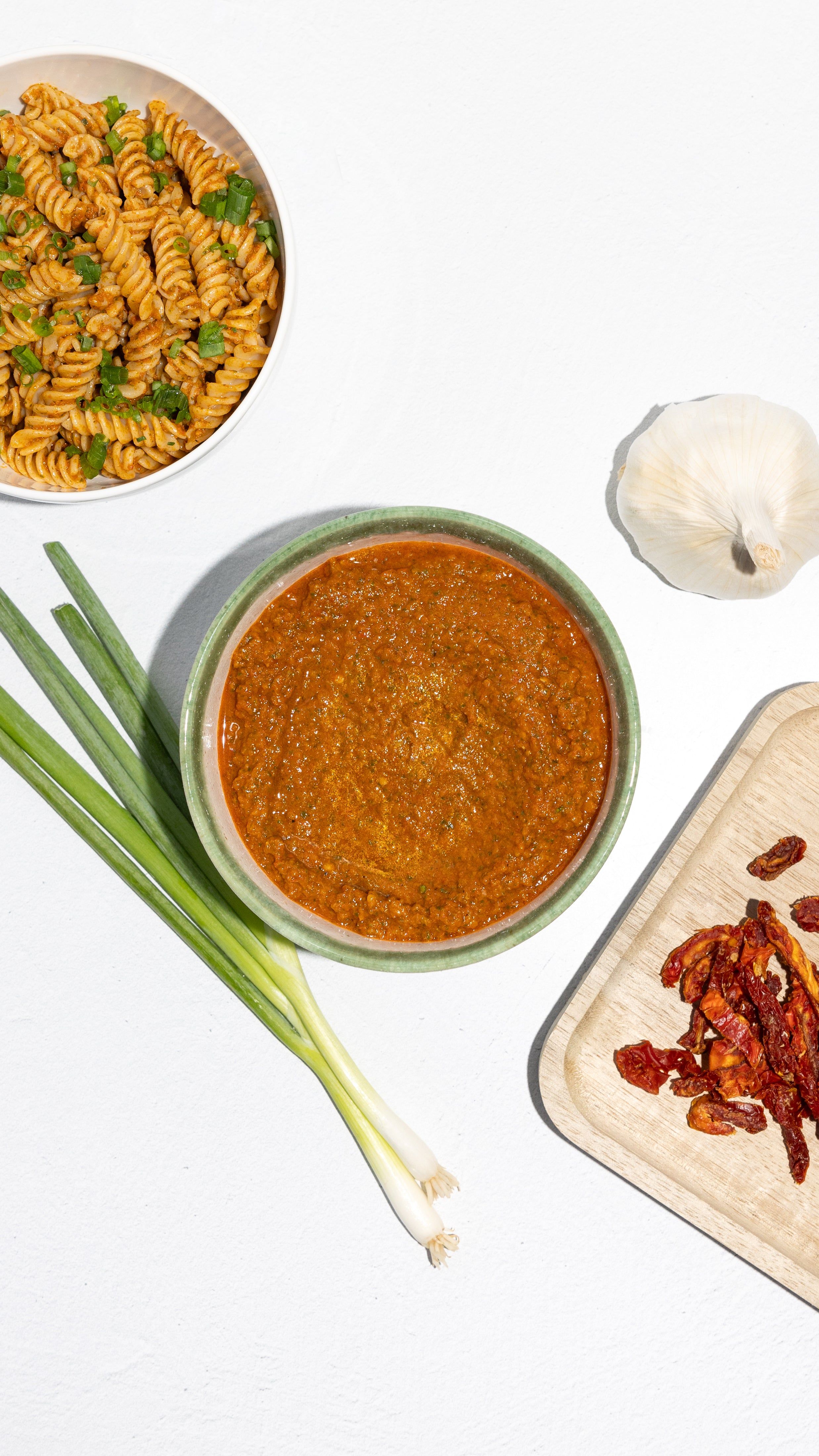 Bowl of sun-dried tomato pesto beside rotini pasta, scallions, garlic, and dried tomatoes.
