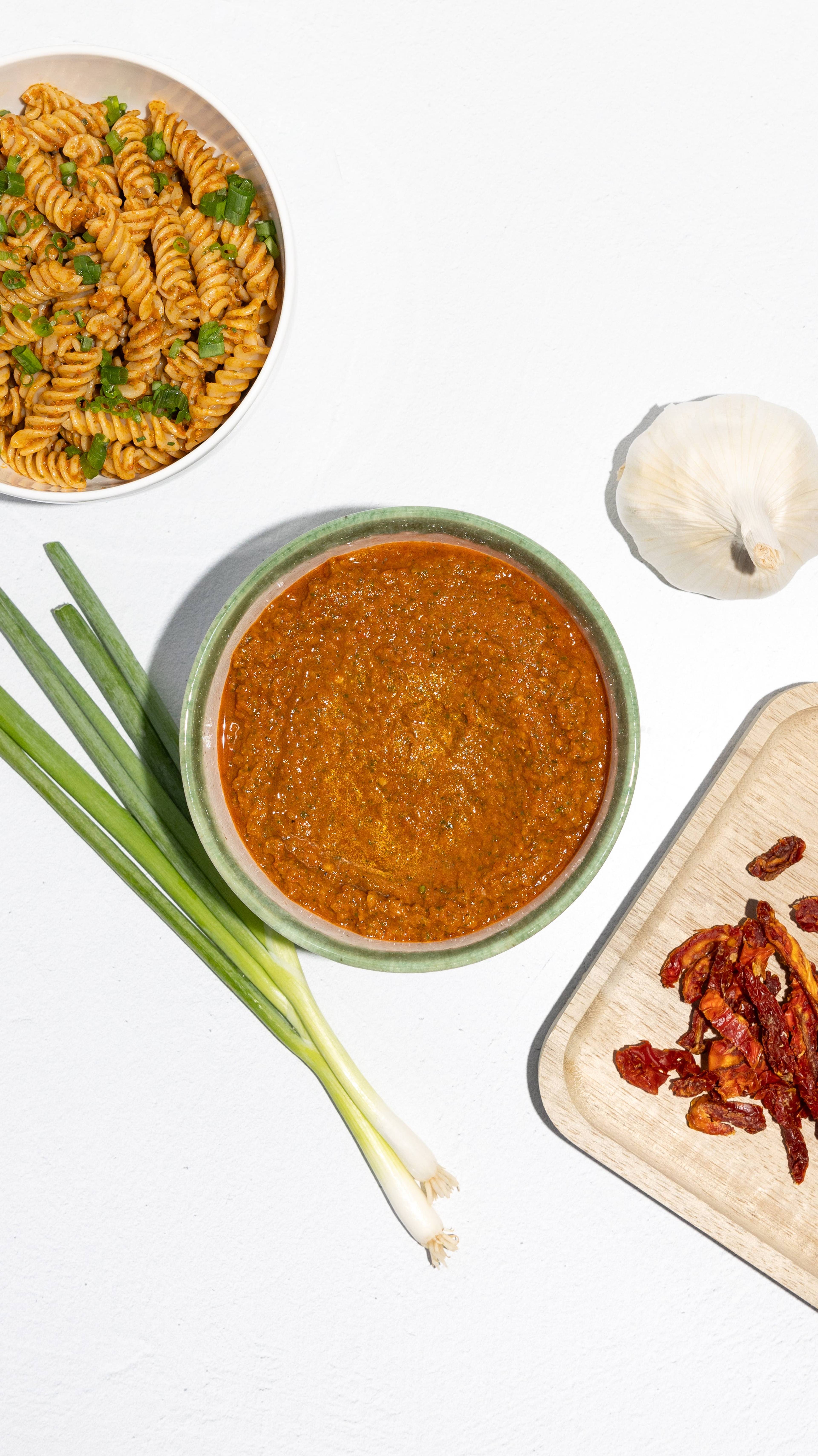 Bowl of sun-dried tomato pesto beside rotini pasta, scallions, garlic, and dried tomatoes.