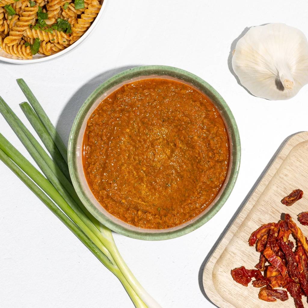 Bowl of sun-dried tomato pesto beside rotini pasta, scallions, garlic, and dried tomatoes.